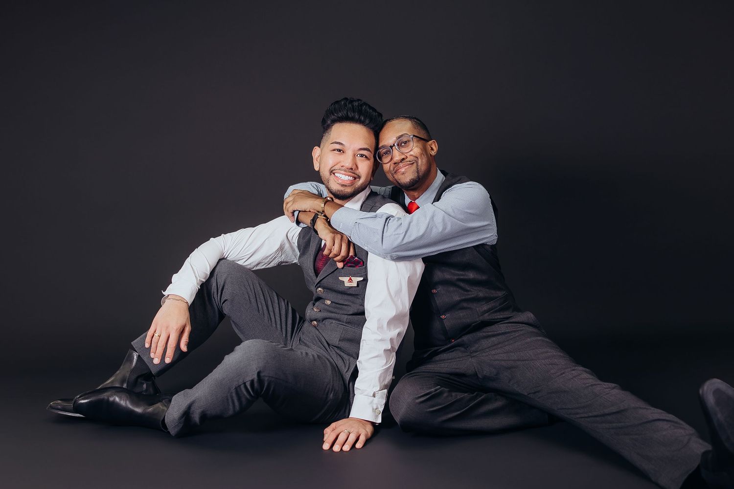Two people in formal attire sitting close together on dark floor against black background.