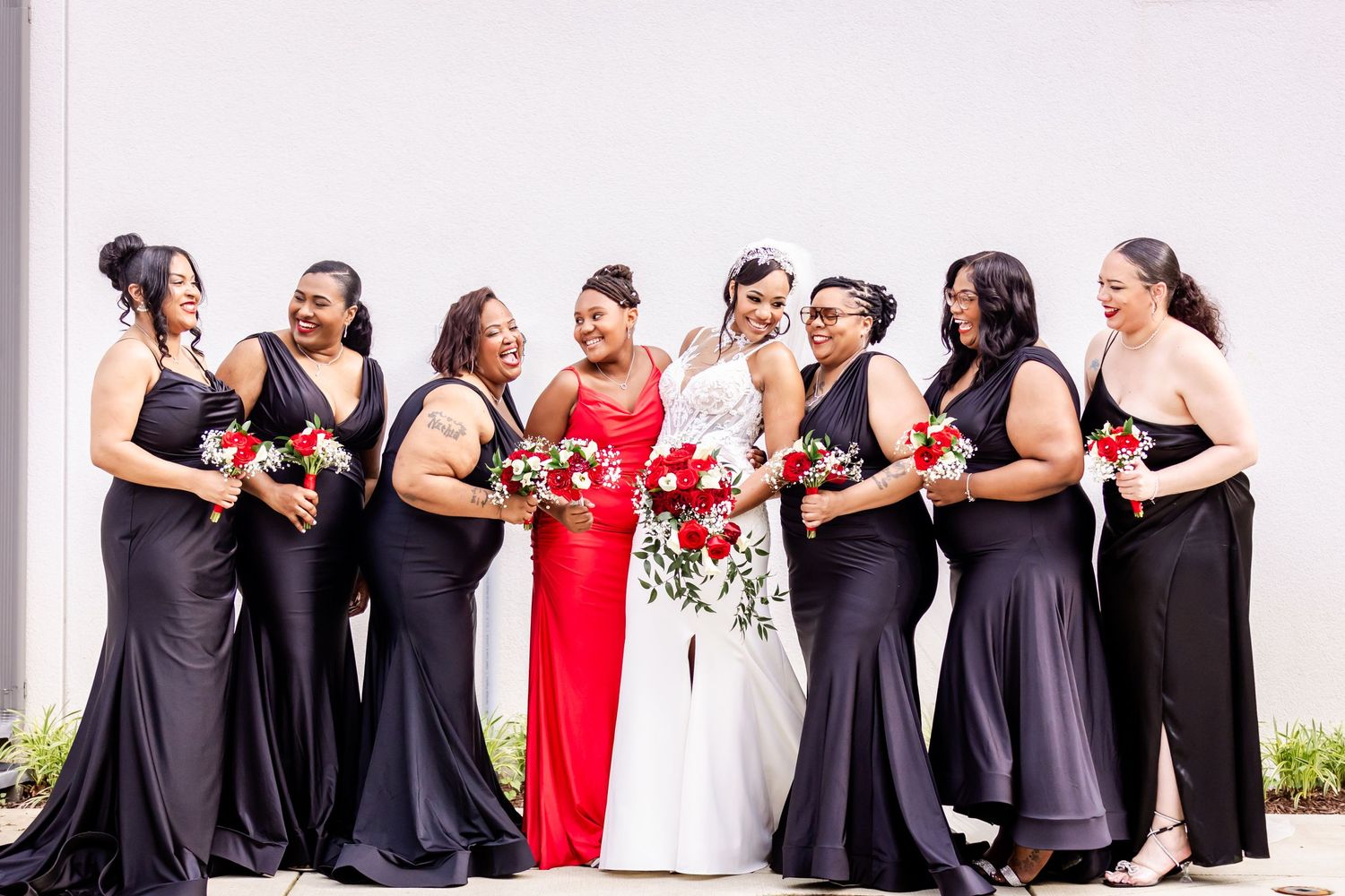 Wedding party in matching navy dresses poses with person in white gown holding red bouquets.