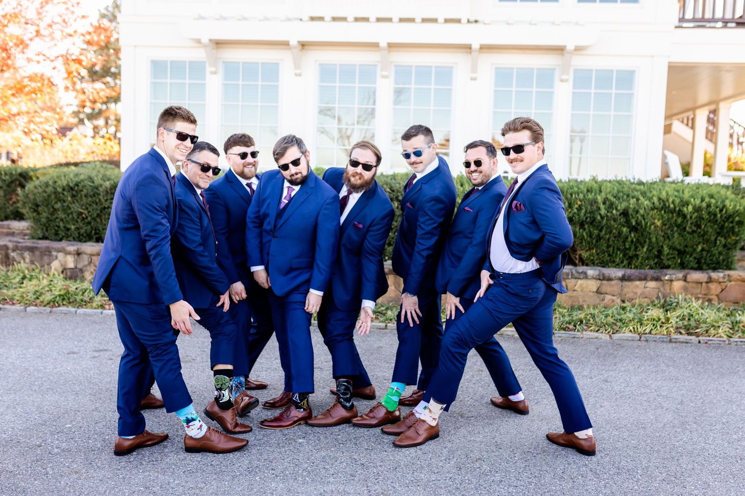 Groomsmen in navy suits strike playful poses wearing sunglasses outside a wedding venue.