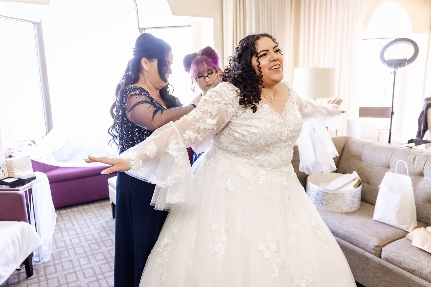 White lace wedding dress being fitted in a hotel room with bridesmaids.