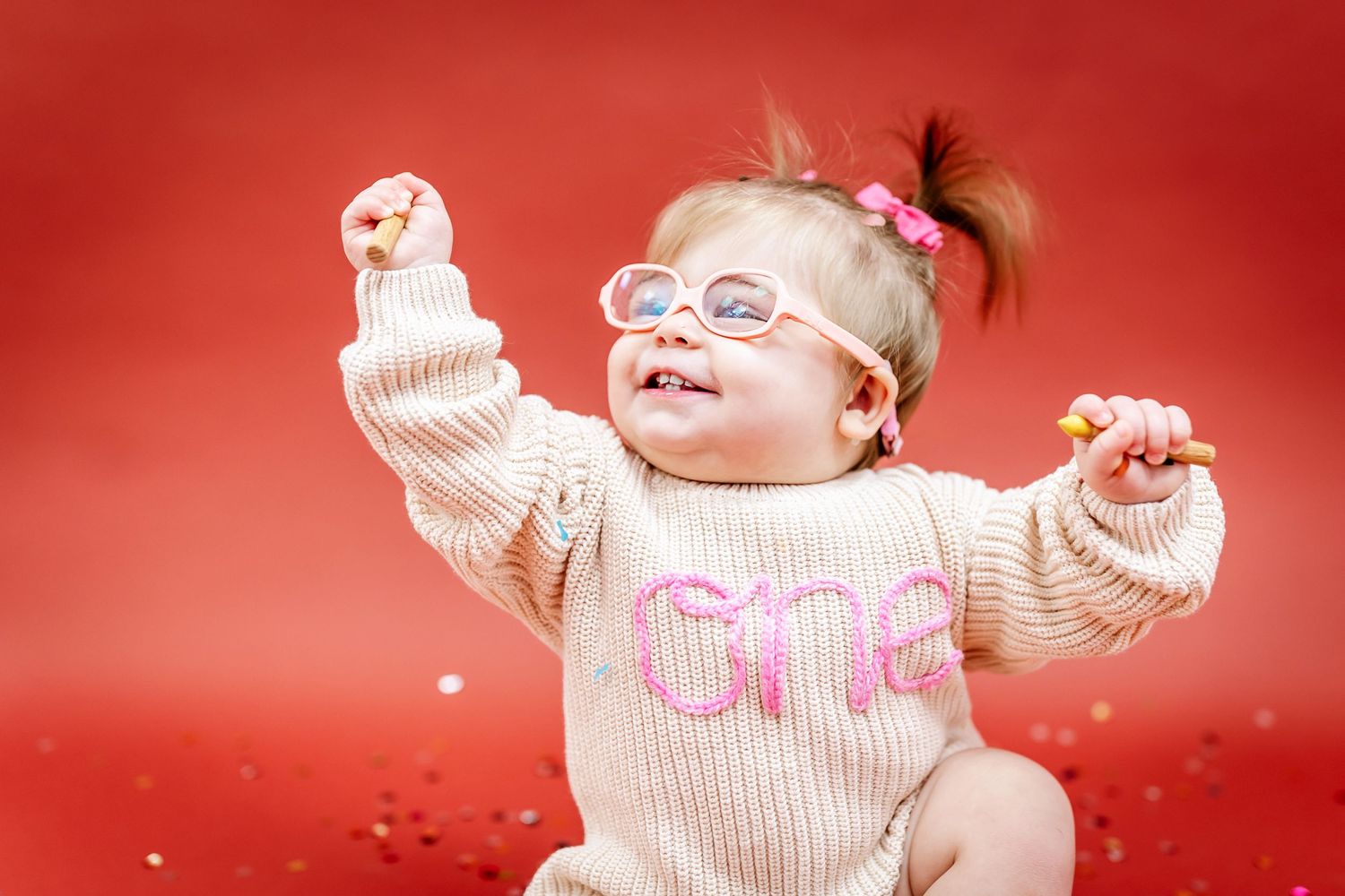 Baby in cream sweater with pink accents shows joy while sitting on red background.