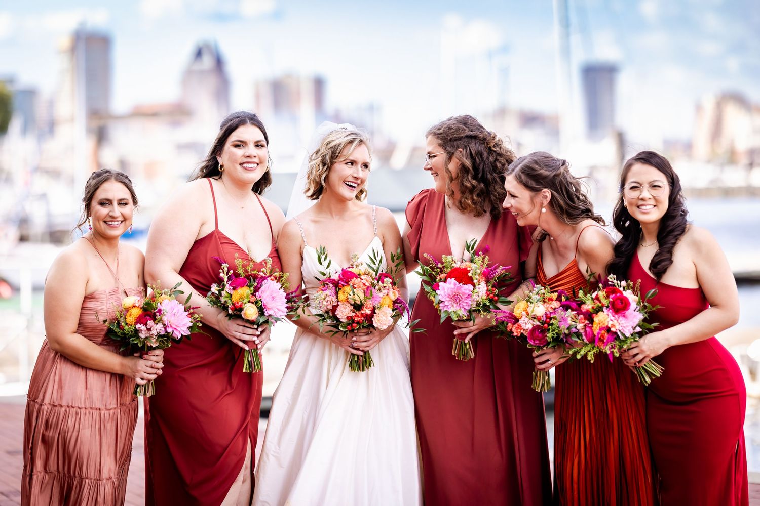 A bridal party in red and blush dresses holding vibrant floral bouquets poses against a city skyline backdrop.