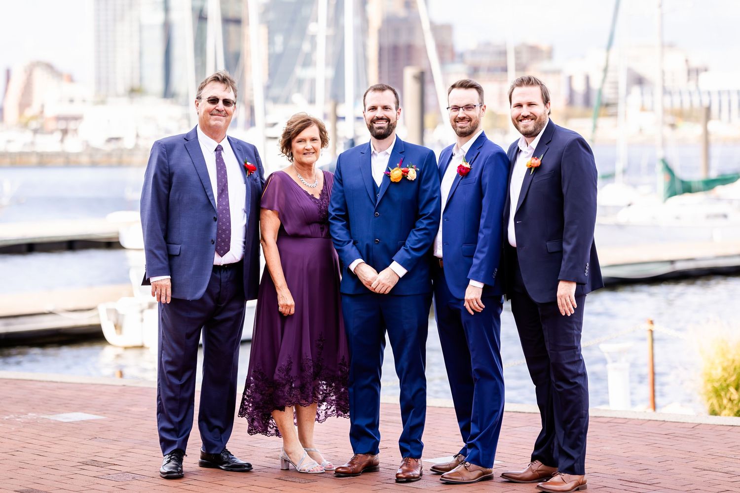 Wedding party poses together on a harborside boardwalk with boats and buildings in the background.