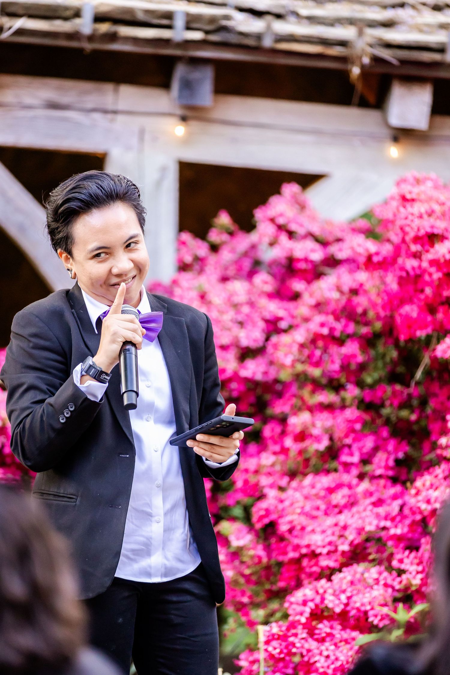 A speaker holds a microphone and phone while standing in front of vibrant pink azalea bushes during an outdoor event.
