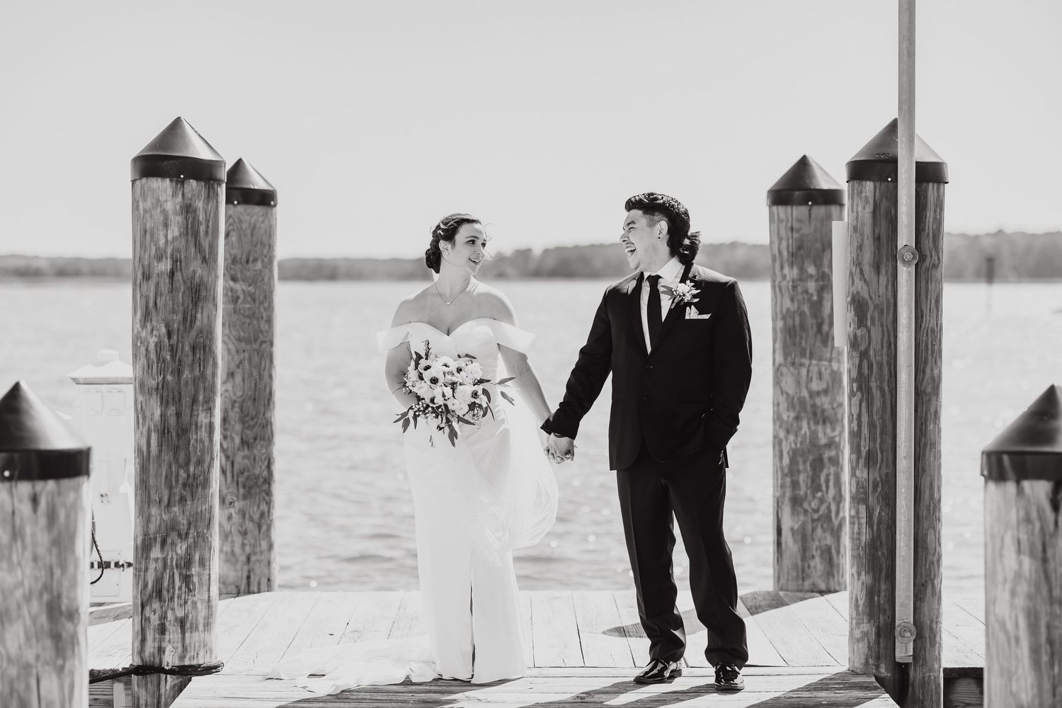 A couple in wedding attire stand hand in hand on a wooden pier overlooking a lake in black and white photography.