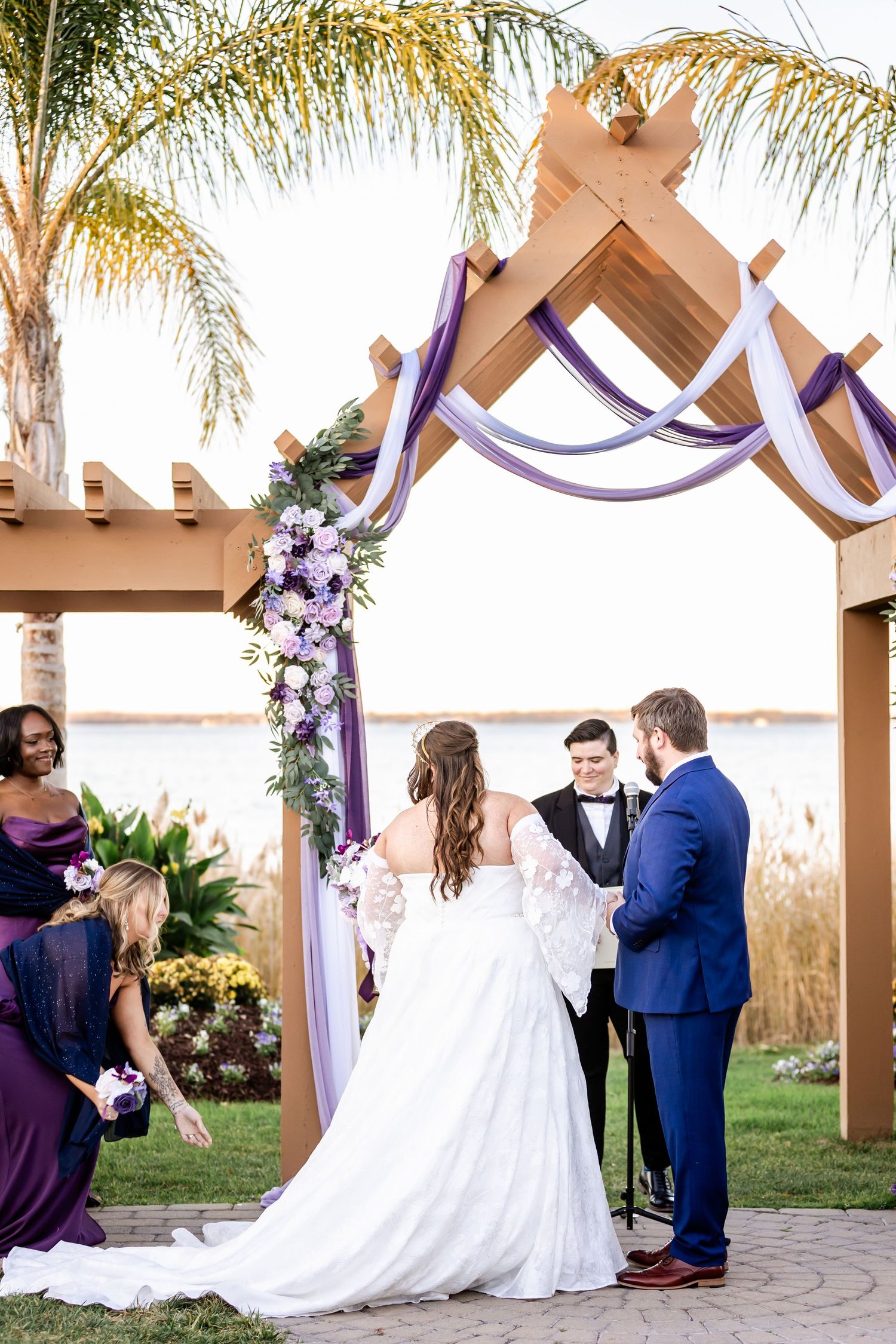 Wedding ceremony under wooden pergola decorated with purple fabric and floral arrangements.