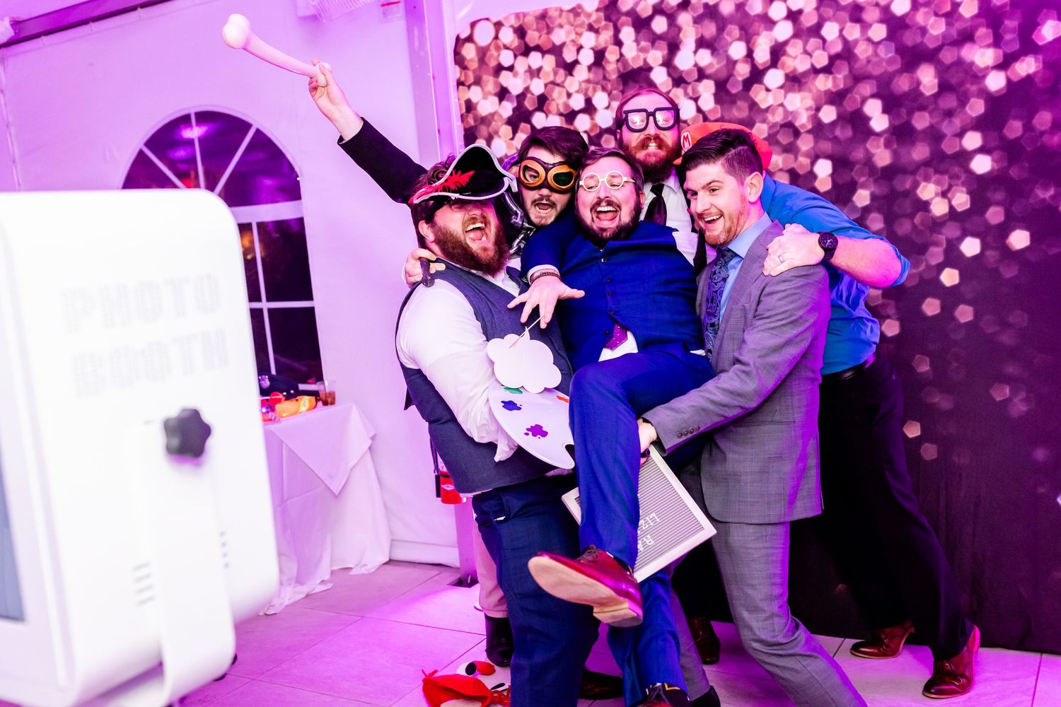 Group celebrating with fun poses under pink lighting at indoor wedding reception.