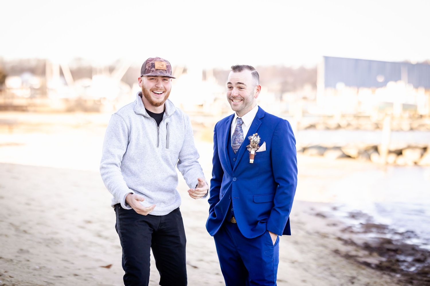 Two friends stand together on a beach, one wearing a blue suit and boutonniere and the other in casual attire.