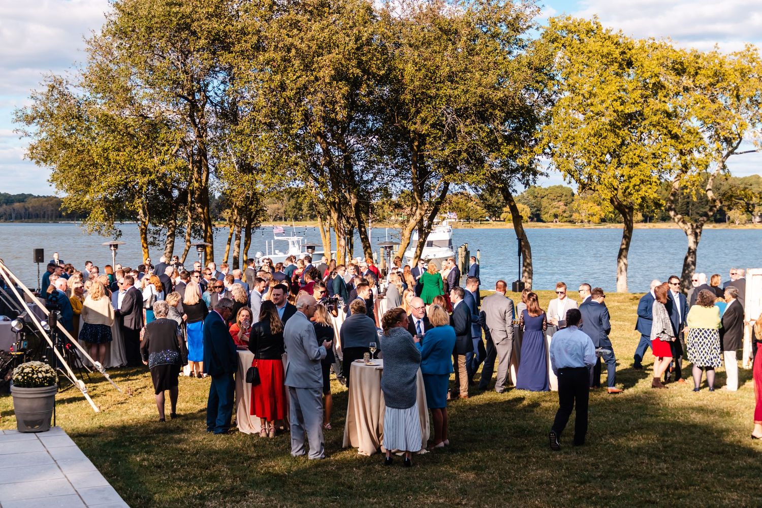 A large outdoor gathering of guests at a lakeside venue with trees during a sunny day.