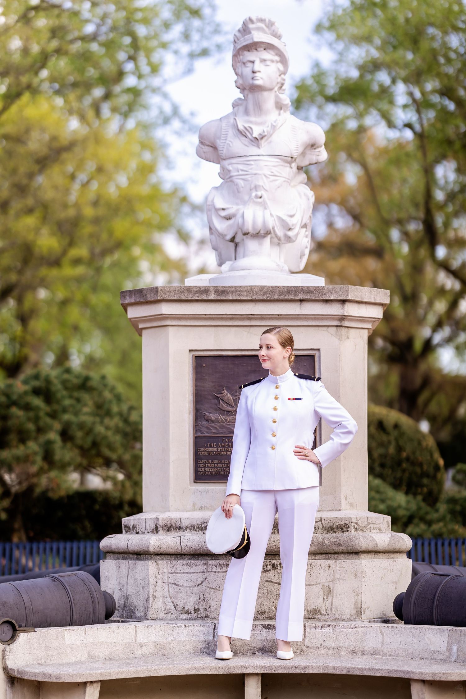 A person in a white naval uniform poses confidently next to a stone monument with a bust and canons in an outdoor setting.