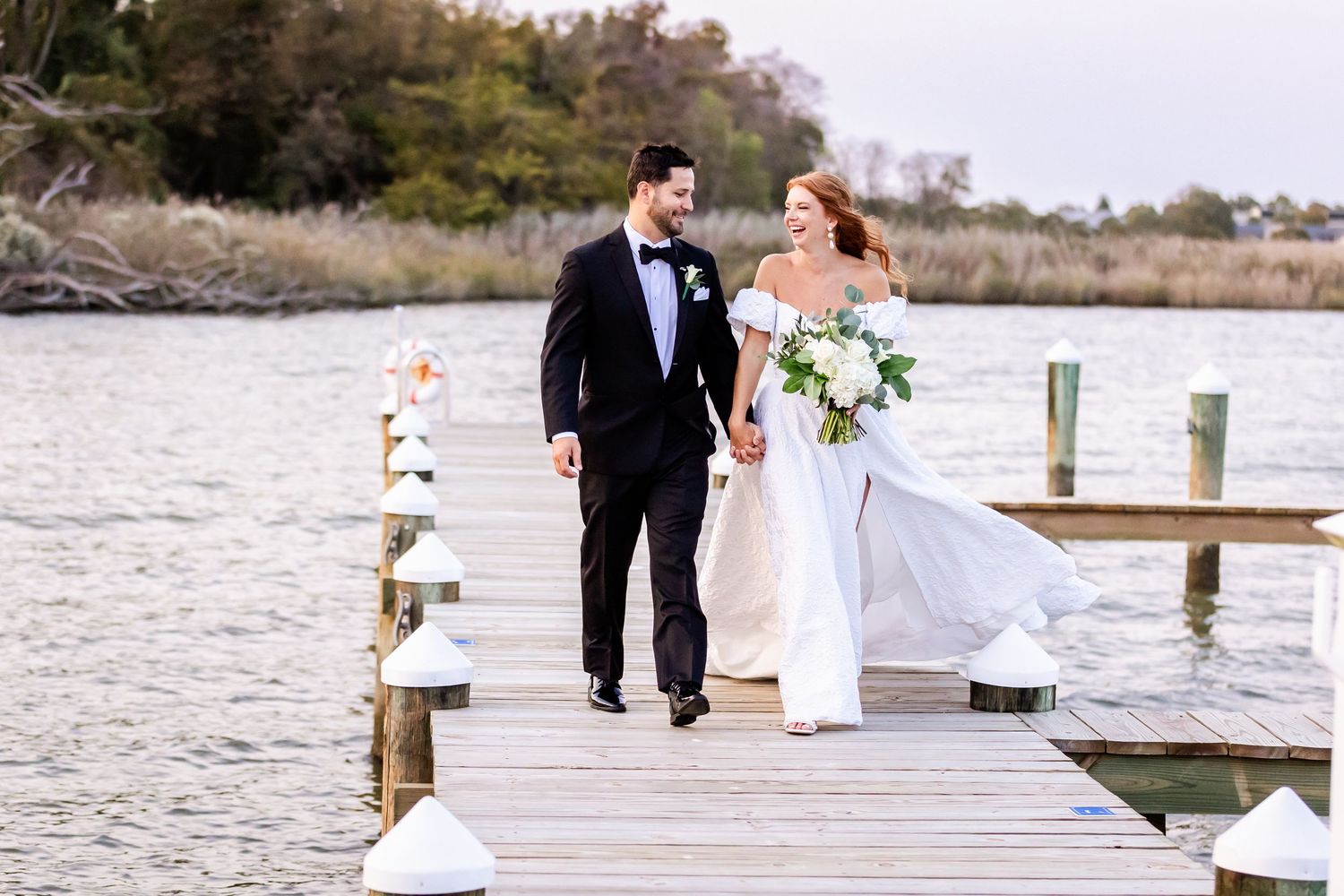A couple walking hand in hand on a wooden dock over water during their outdoor waterfront wedding ceremony.