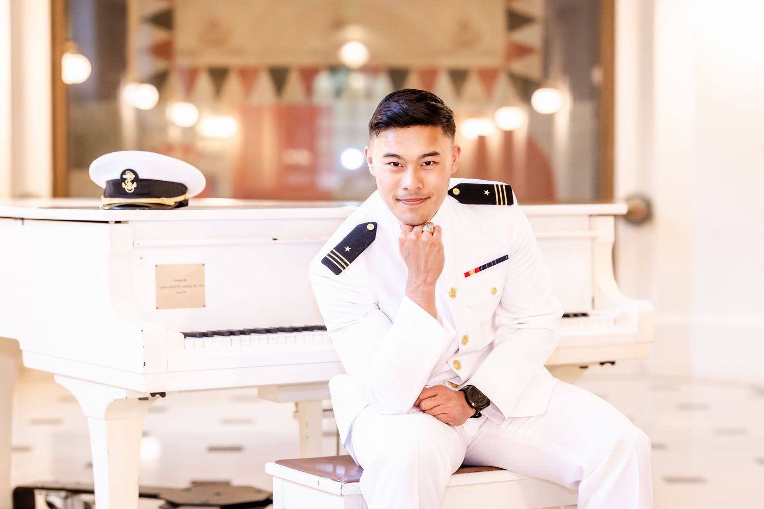 Someone in a crisp white naval uniform poses beside an elegant white grand piano in a formal setting.