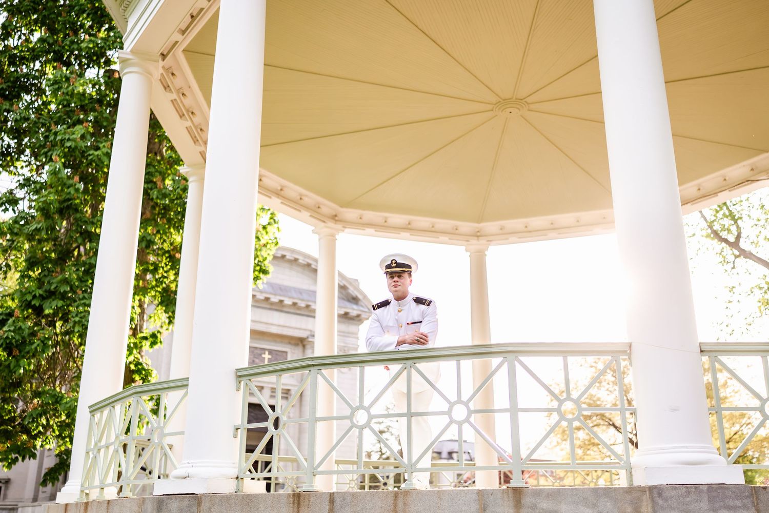 White gazebo with decorative columns and railing on a sunny day surrounded by greenery.