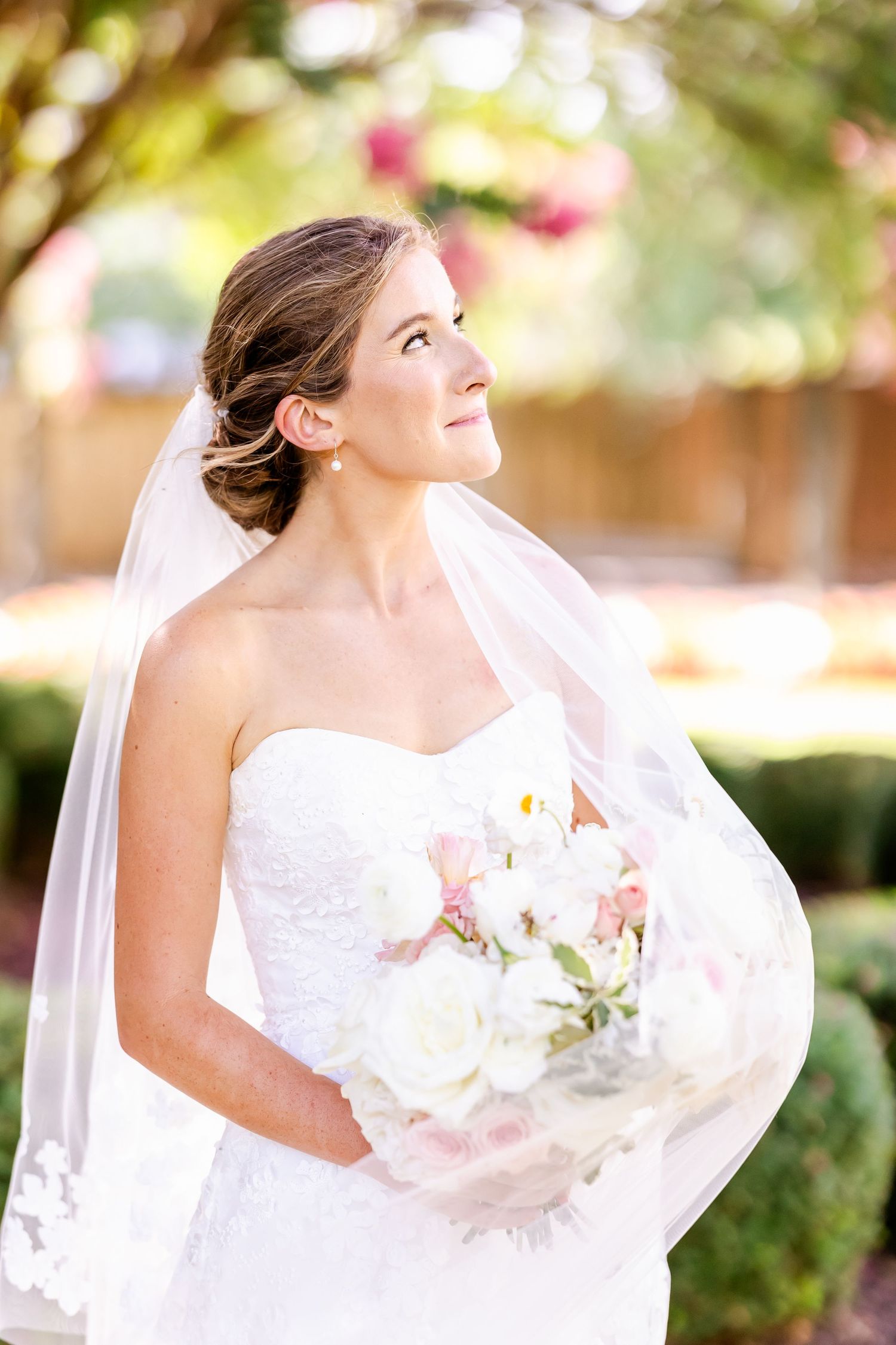 A bride in white wedding dress looks upward while holding a bouquet in an outdoor garden setting.