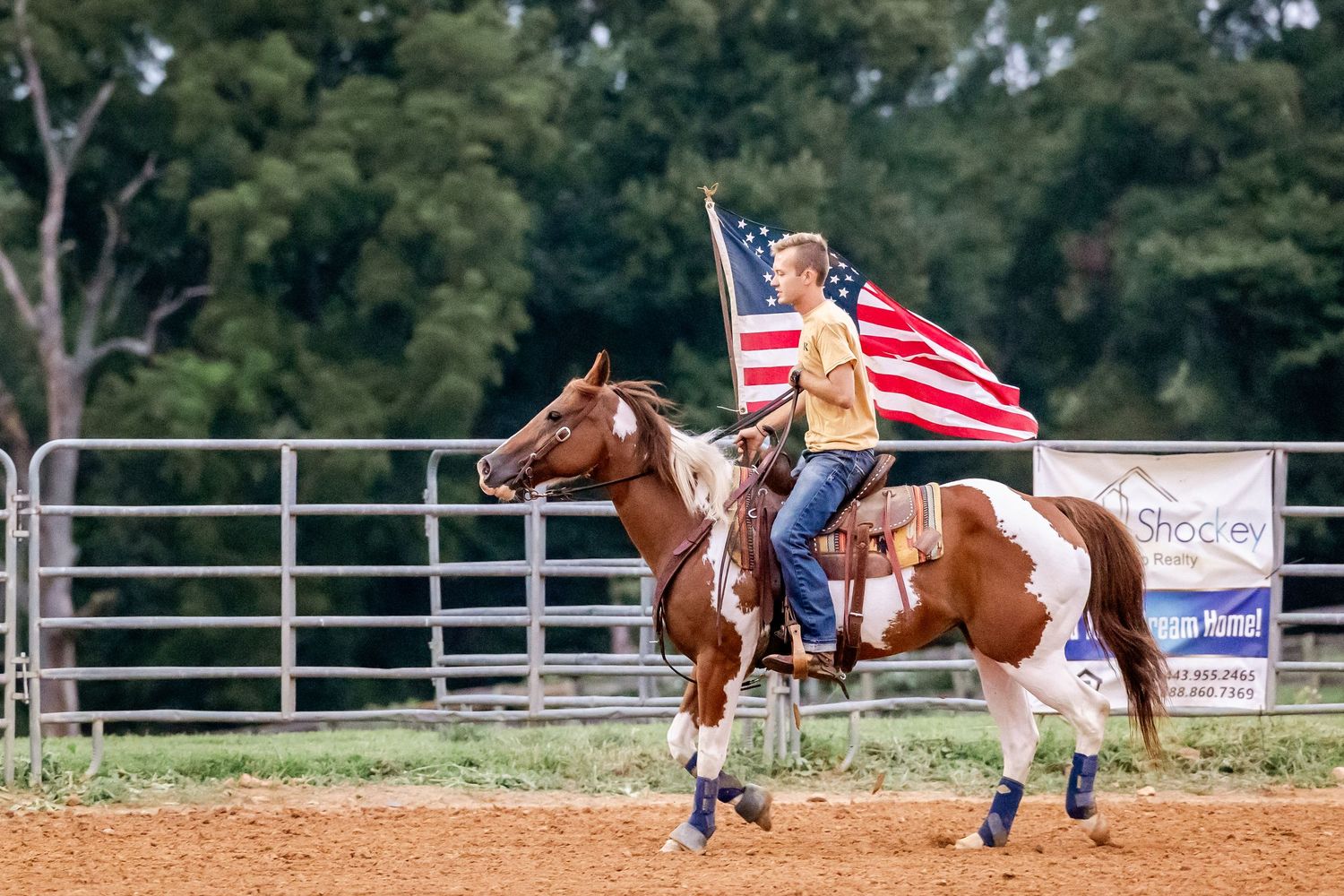 Rider waves American flag while galloping on paint horse during rodeo event.