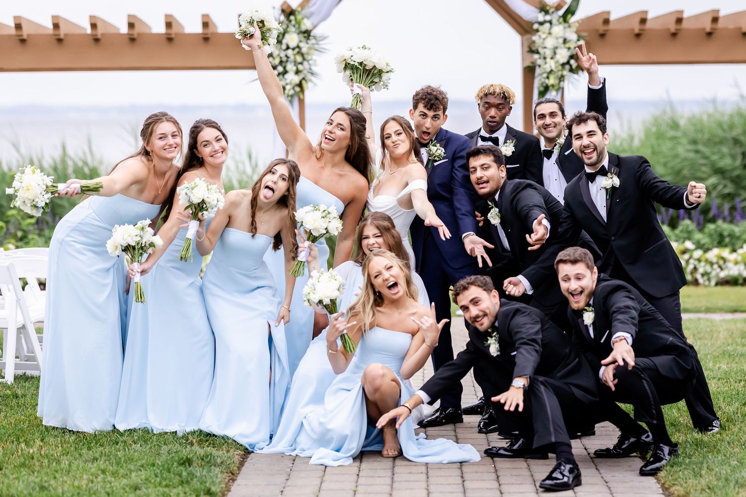 Wedding party in light blue and black attire poses together outdoors for group photos.