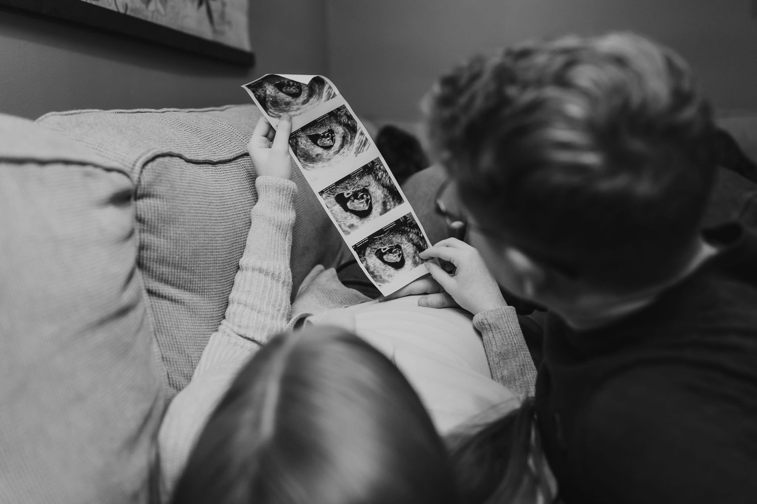 Black and white photo of expectant parents looking at ultrasound pictures on couch.
