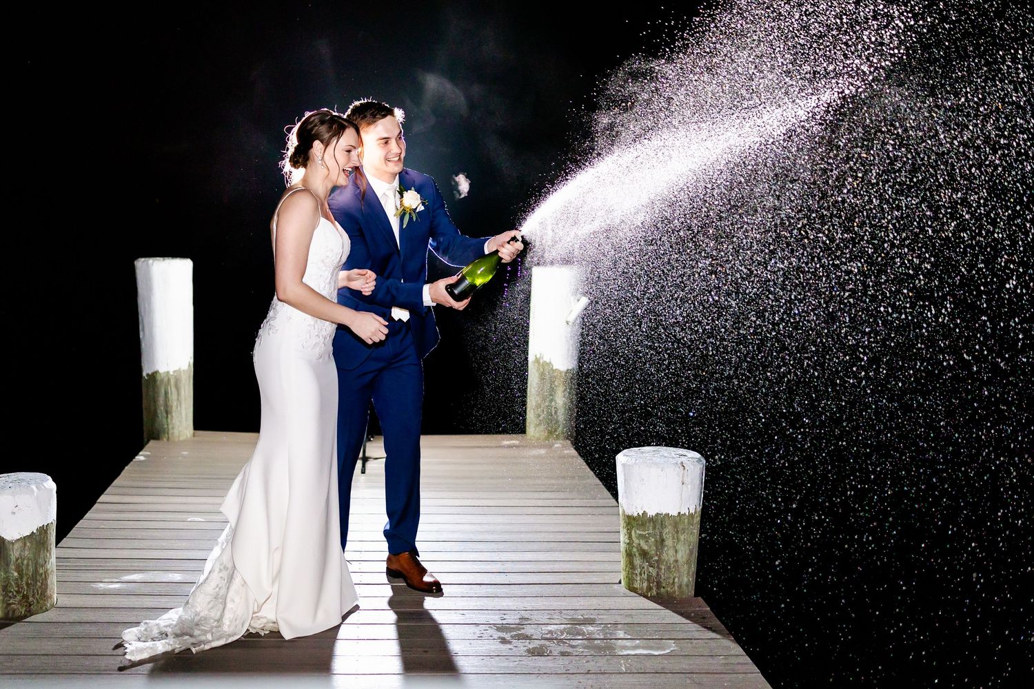 Couple celebrates with champagne spray on wooden dock at night.