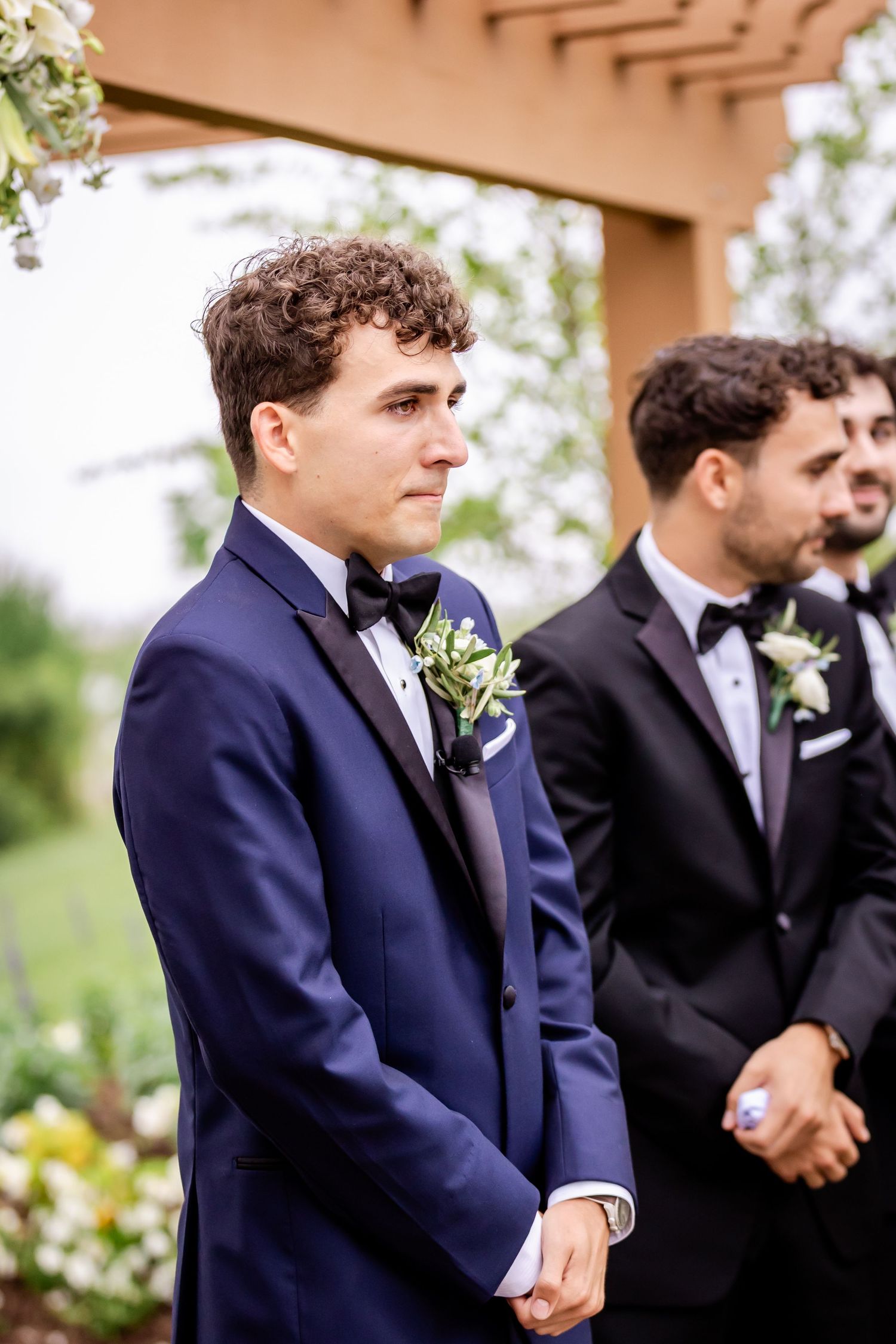 Groomsmen in navy and black suits stand together during an outdoor wedding ceremony.