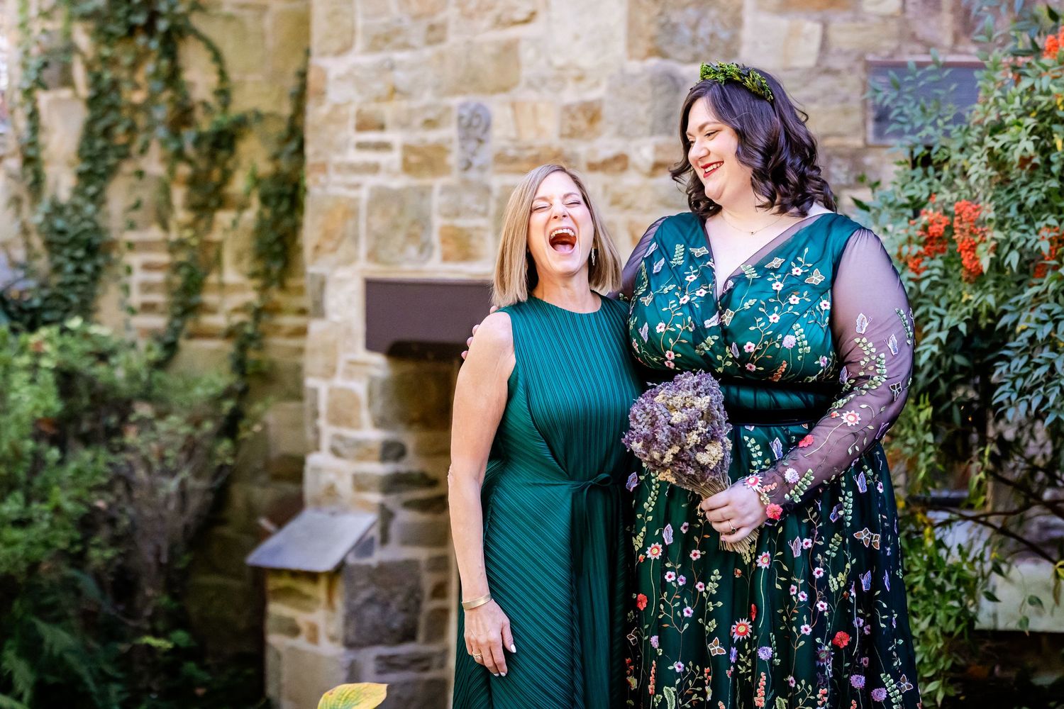 Two women in elegant dresses share an exciting moment outdoors against a stone wall backdrop.