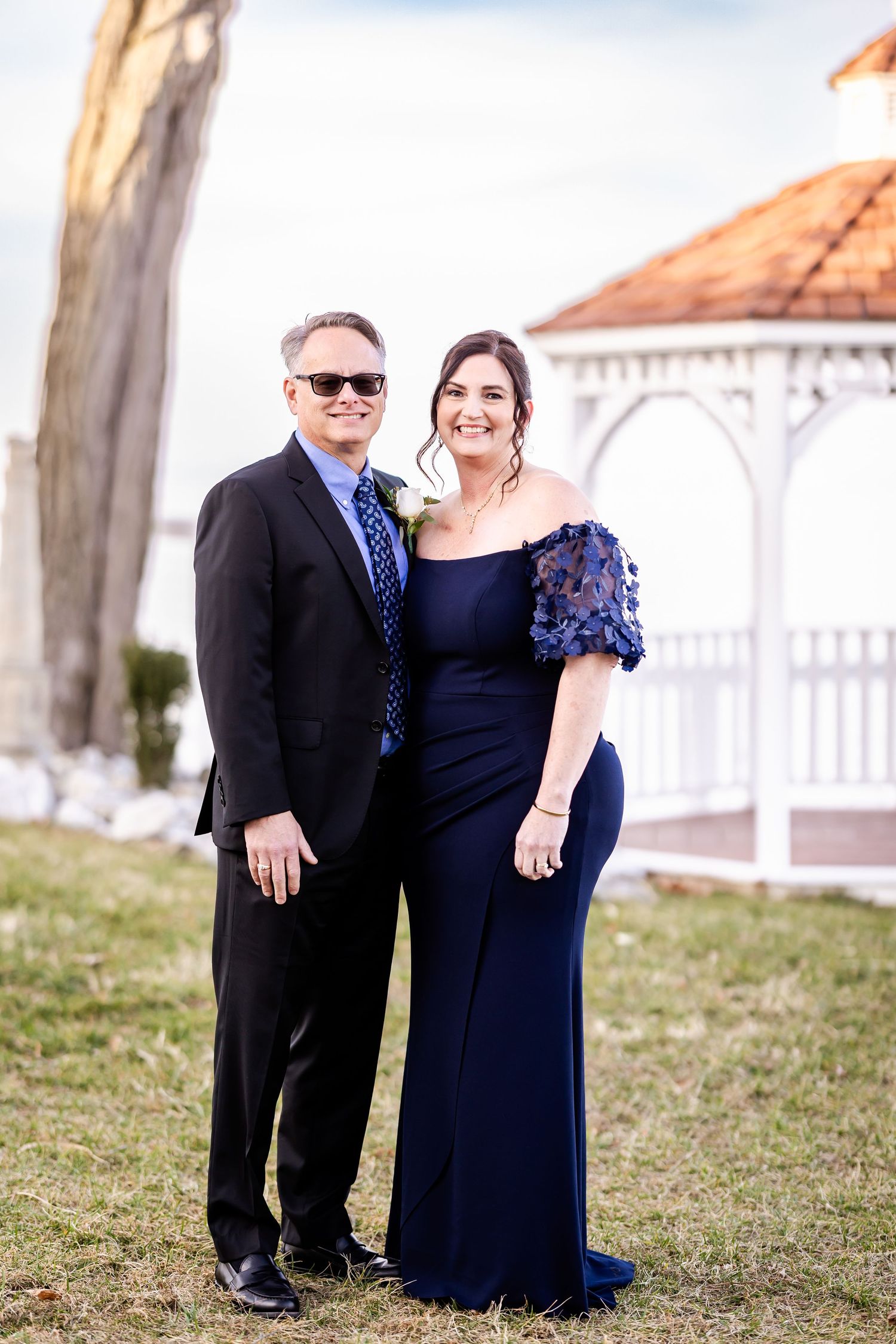 A couple in formal navy blue attire poses together outdoors near a white gazebo.