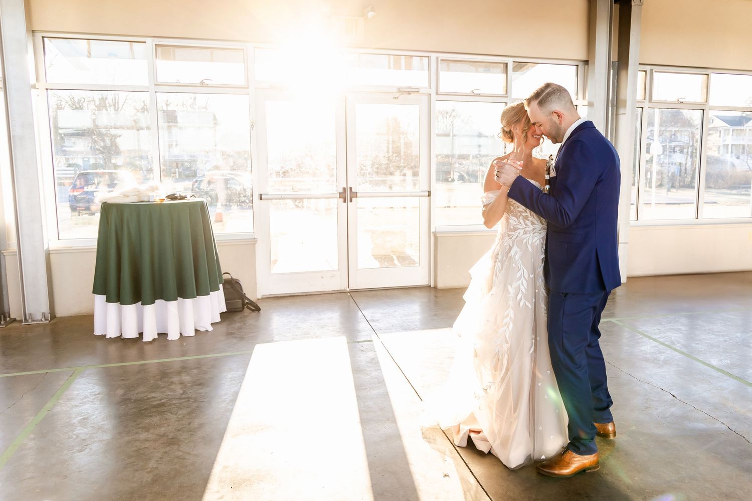 A couple shares their first dance together in a sunlit room with floor-to-ceiling windows during their wedding celebration.
