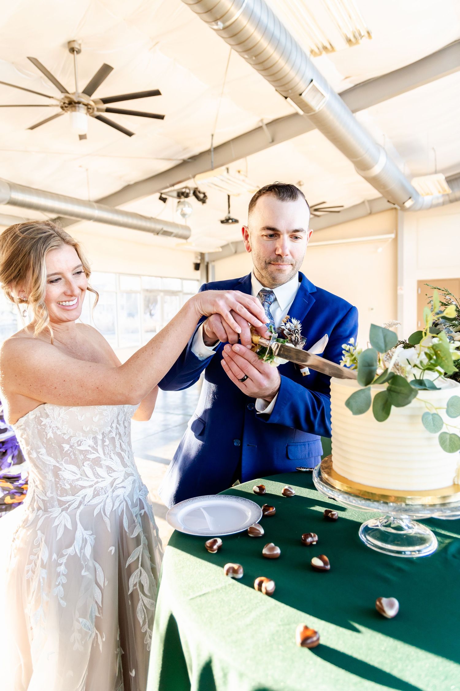 A couple cuts their white wedding cake together on a green tablecloth during their reception celebration.