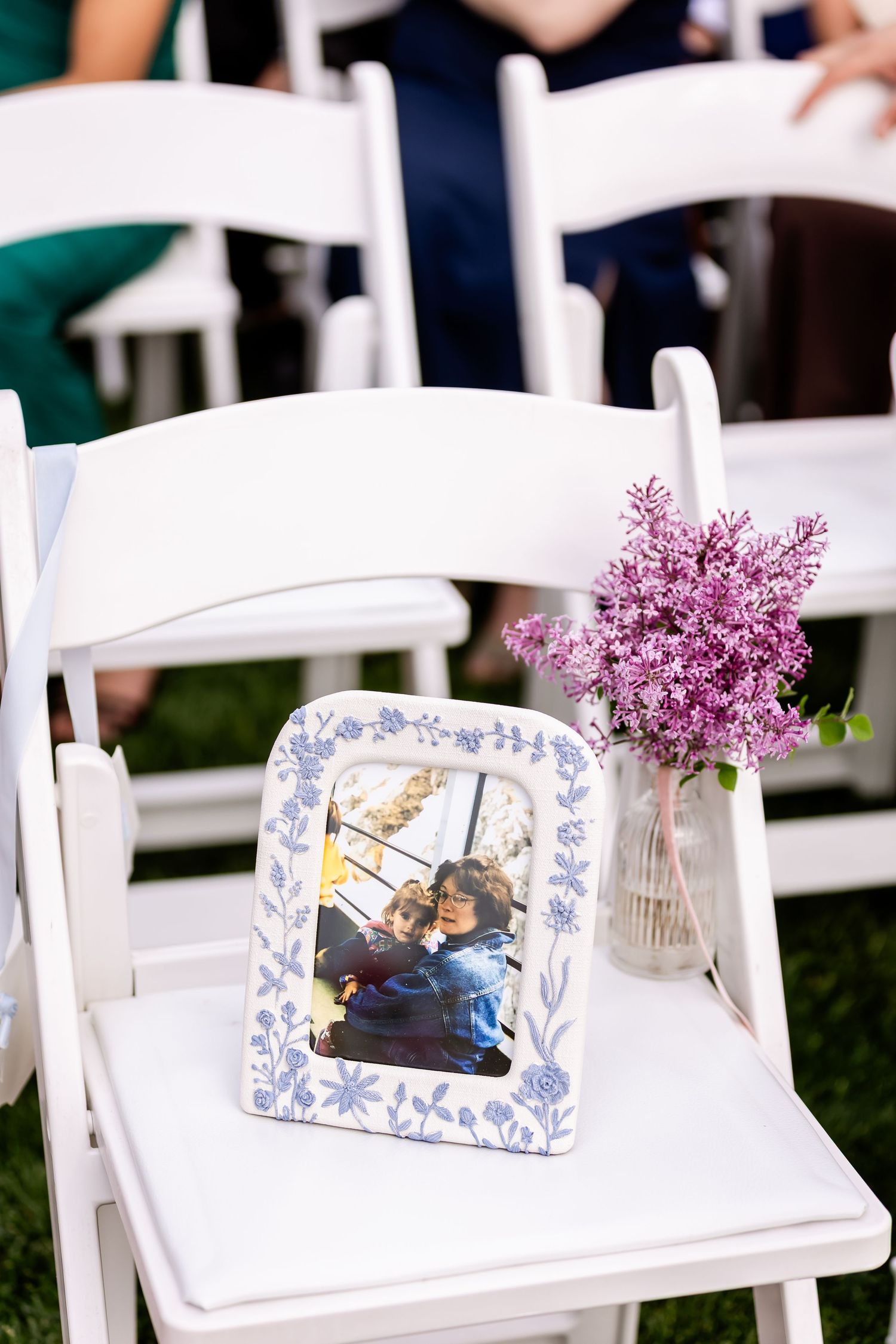 A framed photo and purple lilac flowers decorate a white folding chair at a wedding ceremony.