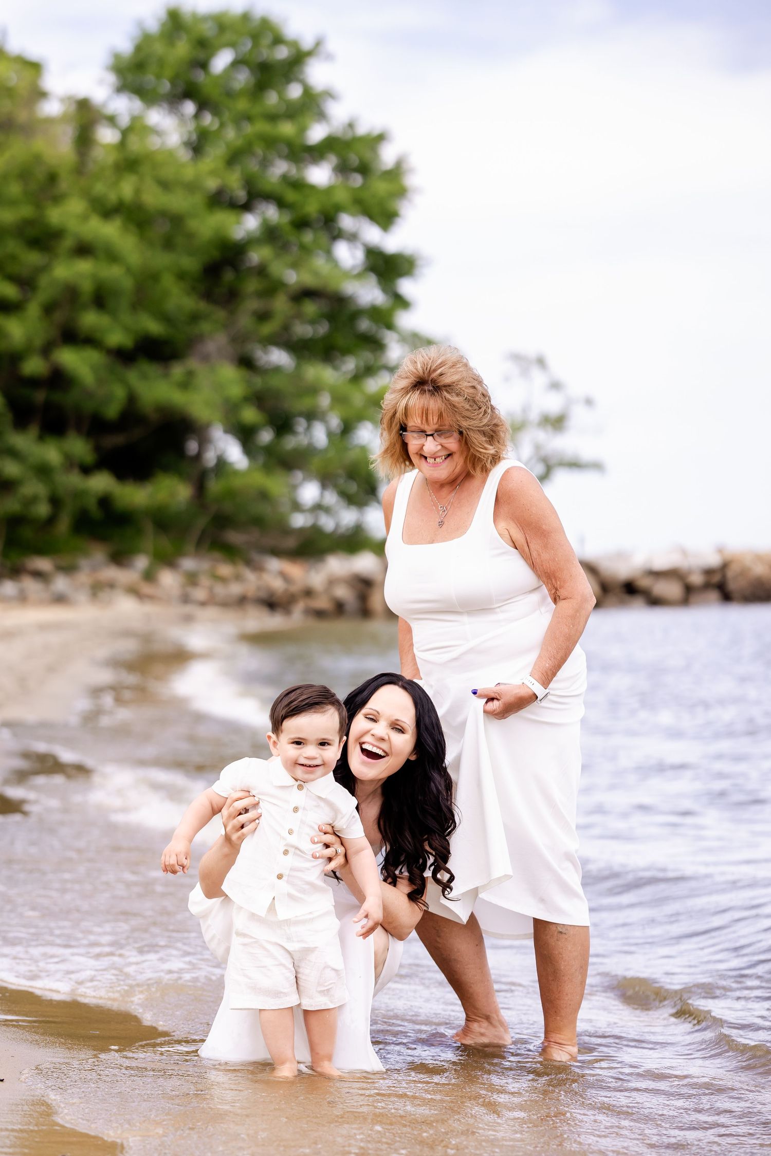 Three generations share joyful moments playing in shallow beach waters while wearing white outfits.