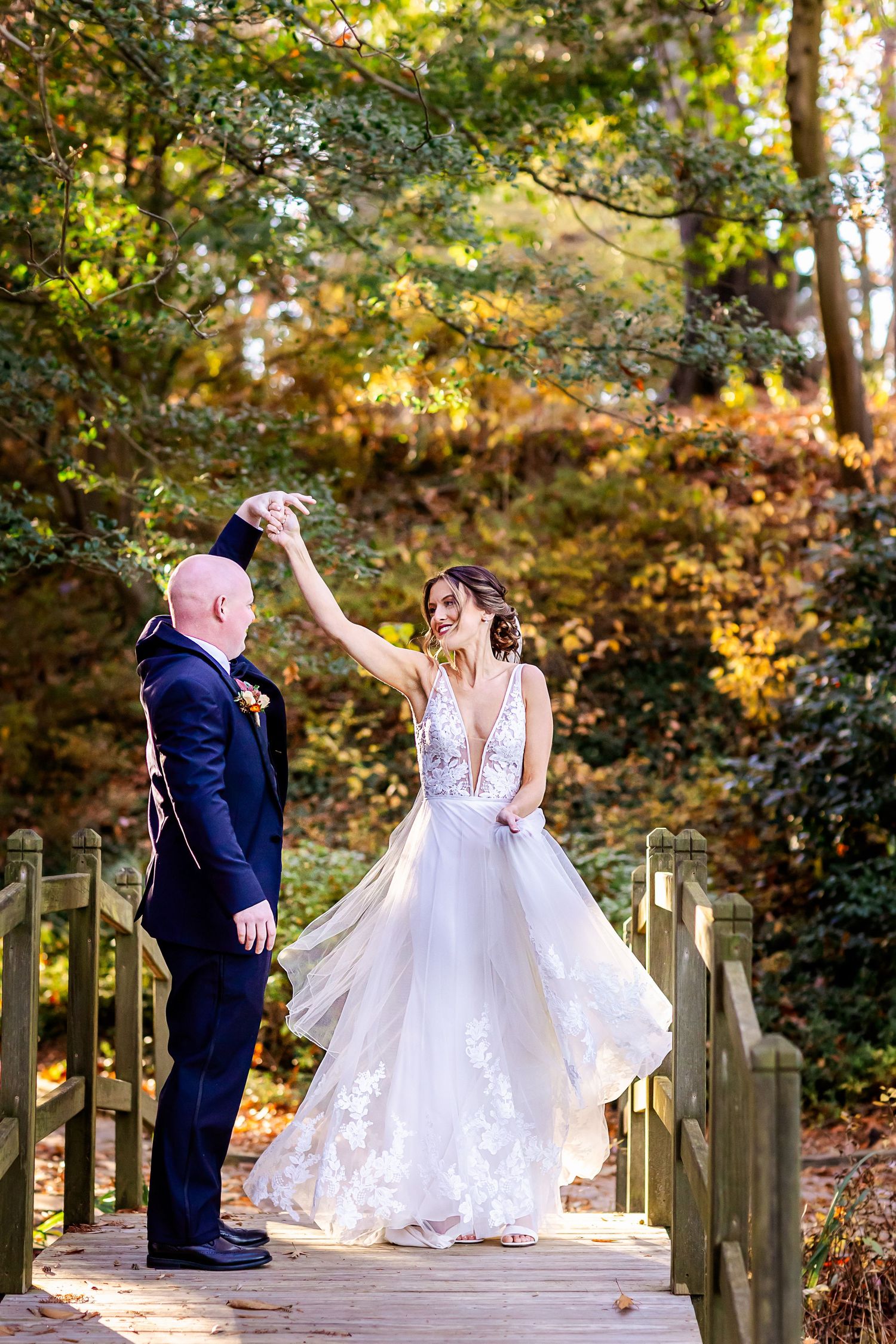 A couple dances together on a wooden footbridge surrounded by autumn foliage in a romantic outdoor setting.