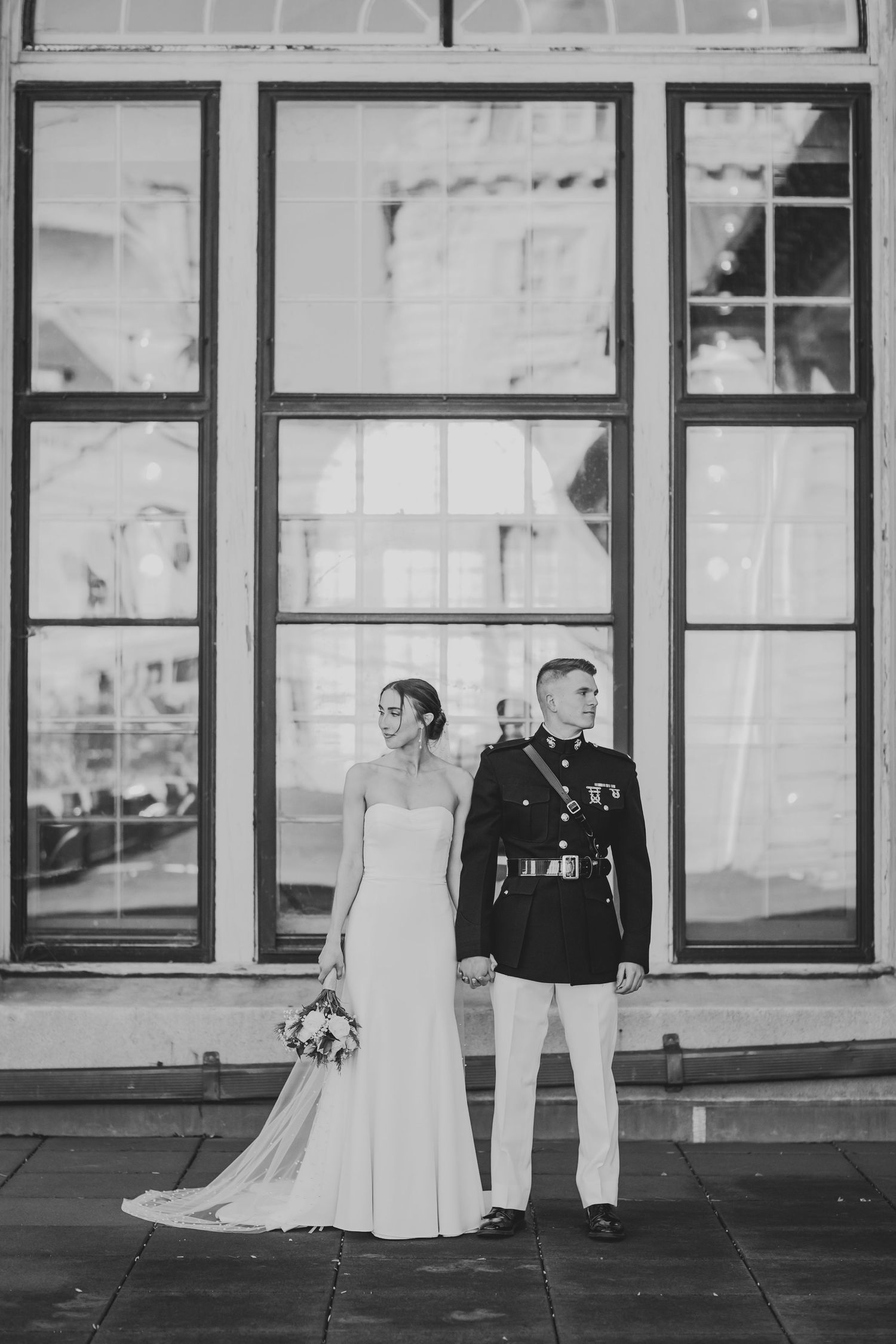 Black and white wedding photo of a couple in formal attire standing in front of large windows with architectural framing.