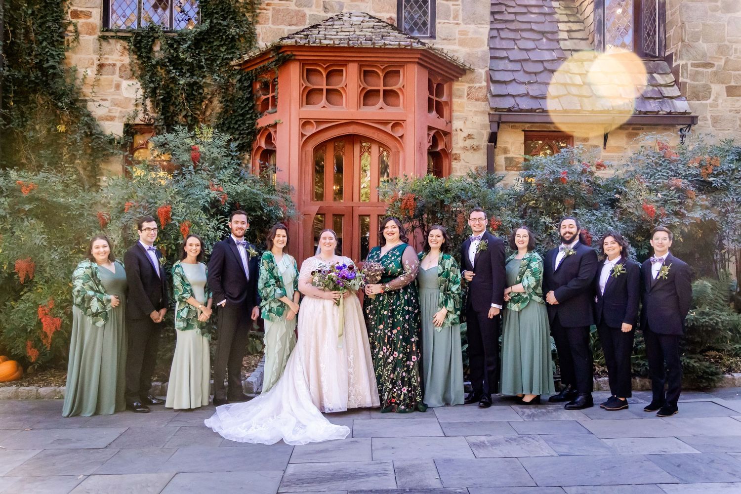 Wedding party in formal attire poses in front of elegant stone church with ornate doorway.