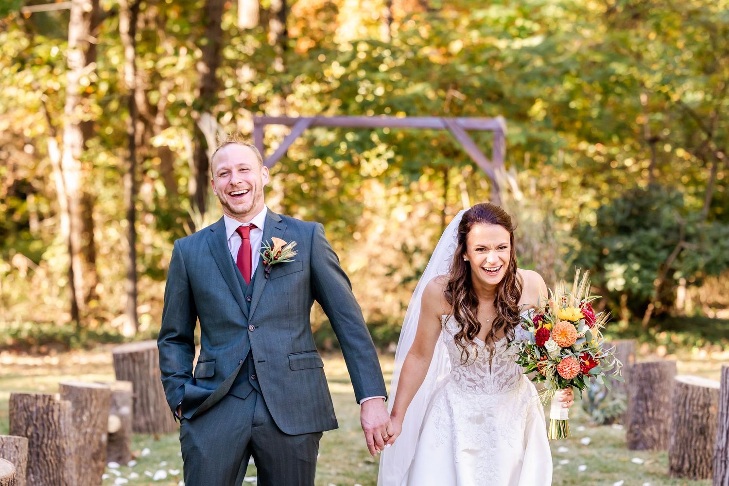 A newlywed couple walks together in a wooded outdoor wedding venue with rustic wooden posts.