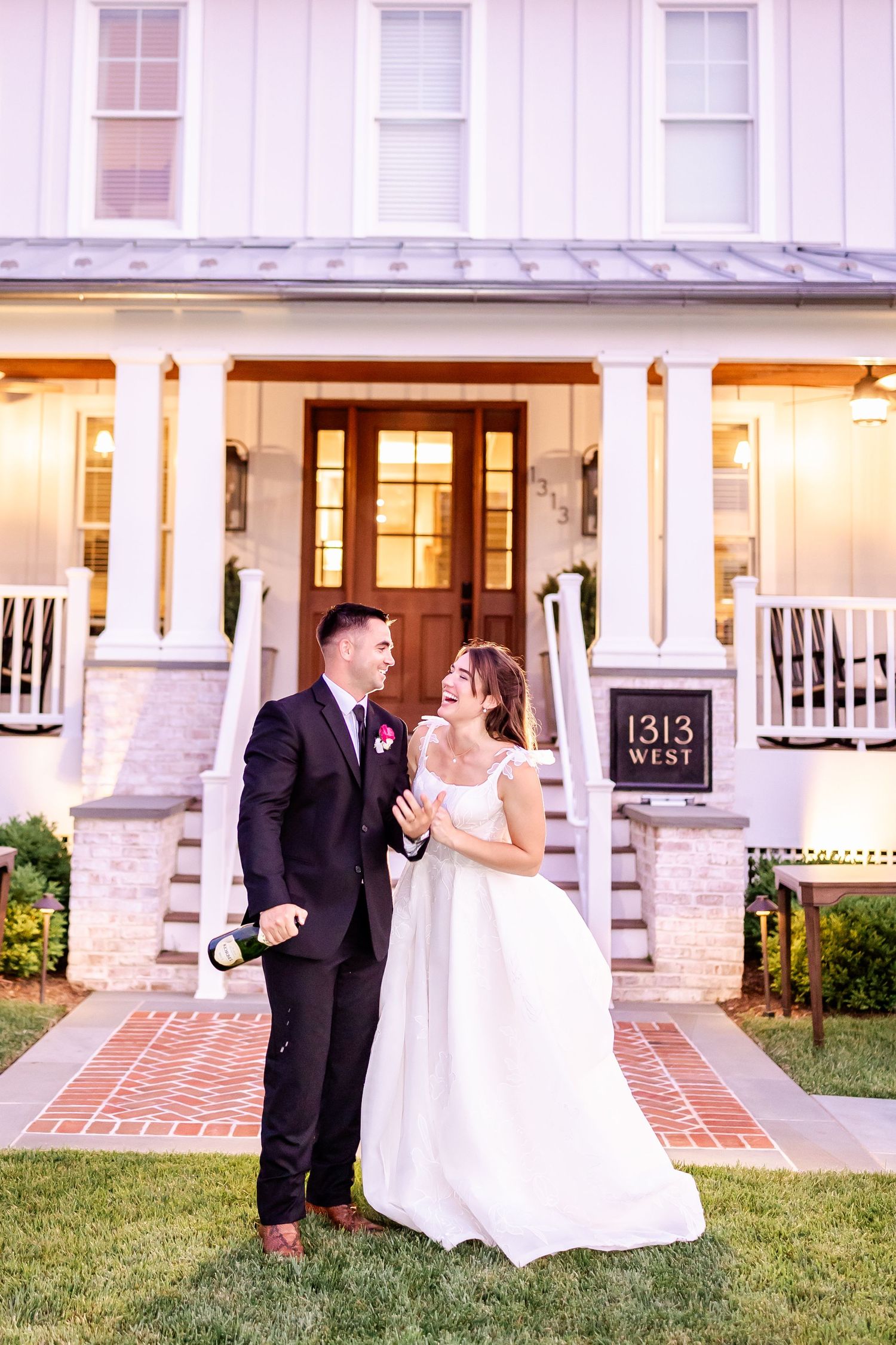 Newlywed couple shares romantic moments in front of a white colonial house with brick pathway at sunset.