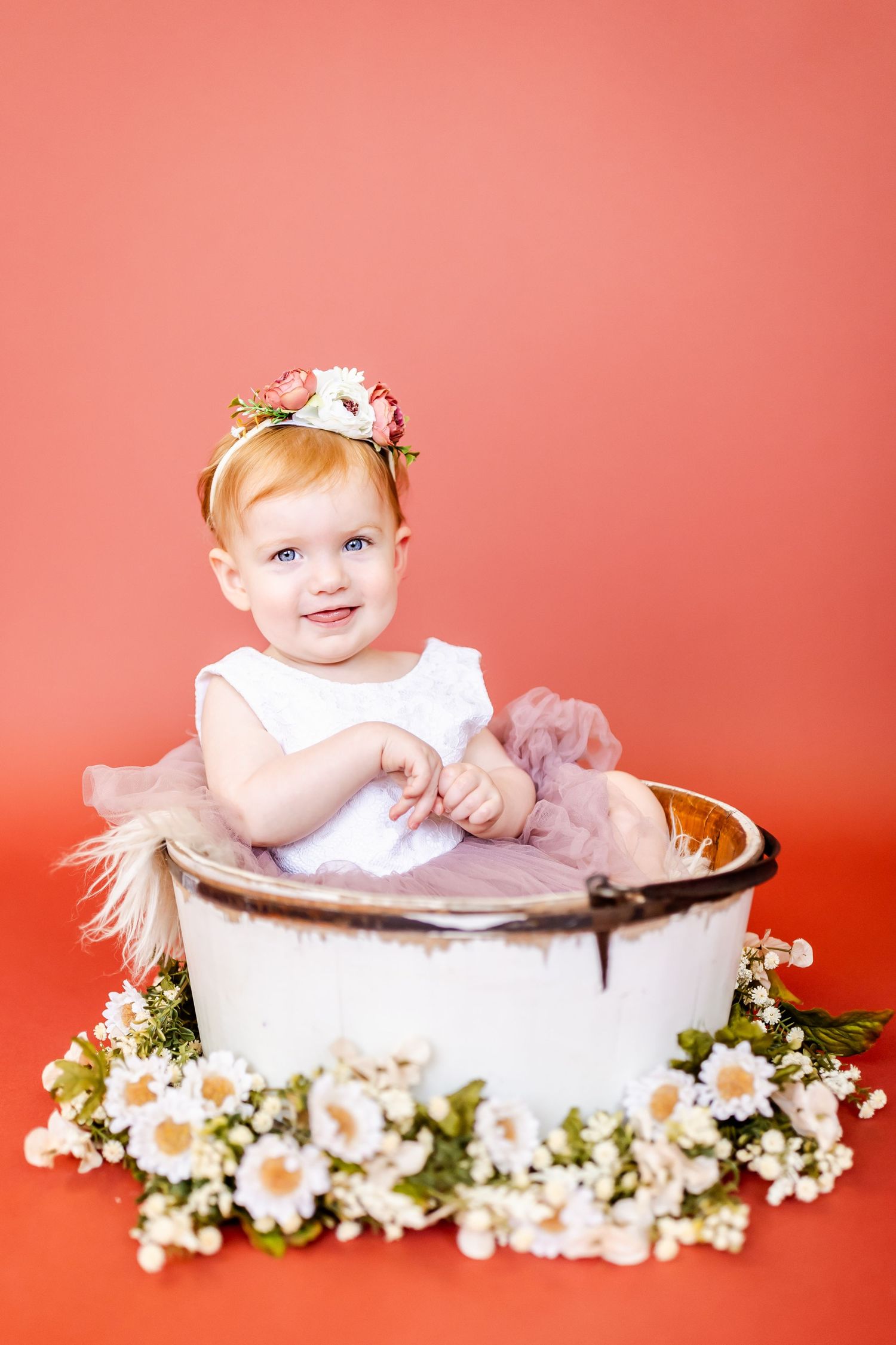 Baby girl sitting in vintage basin surrounded by delicate white flowers against coral background.