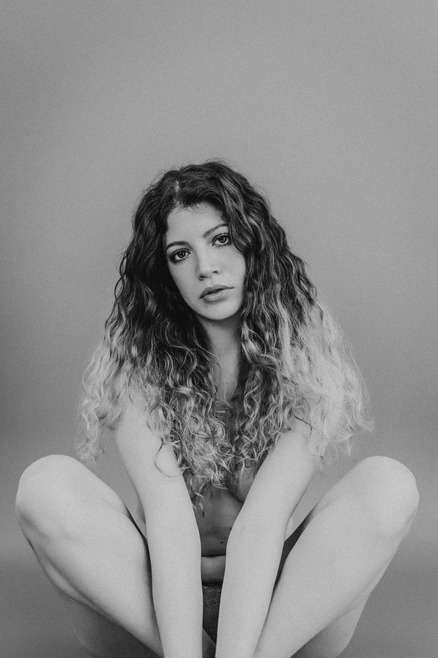 Black and white artistic portrait of a person with long curly ombre hair sitting in a minimalist pose against plain backdrop.