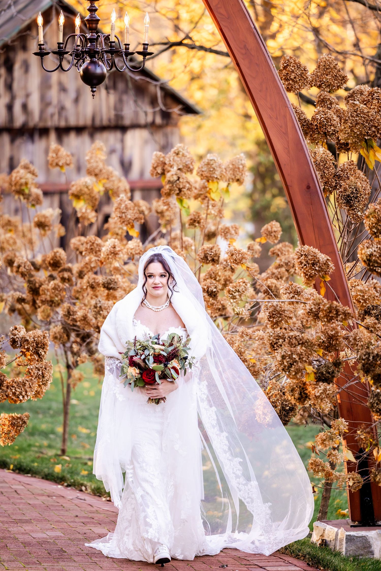 A bride in white lace holds a burgundy bouquet under a wooden arch with autumn foliage and chandelier.