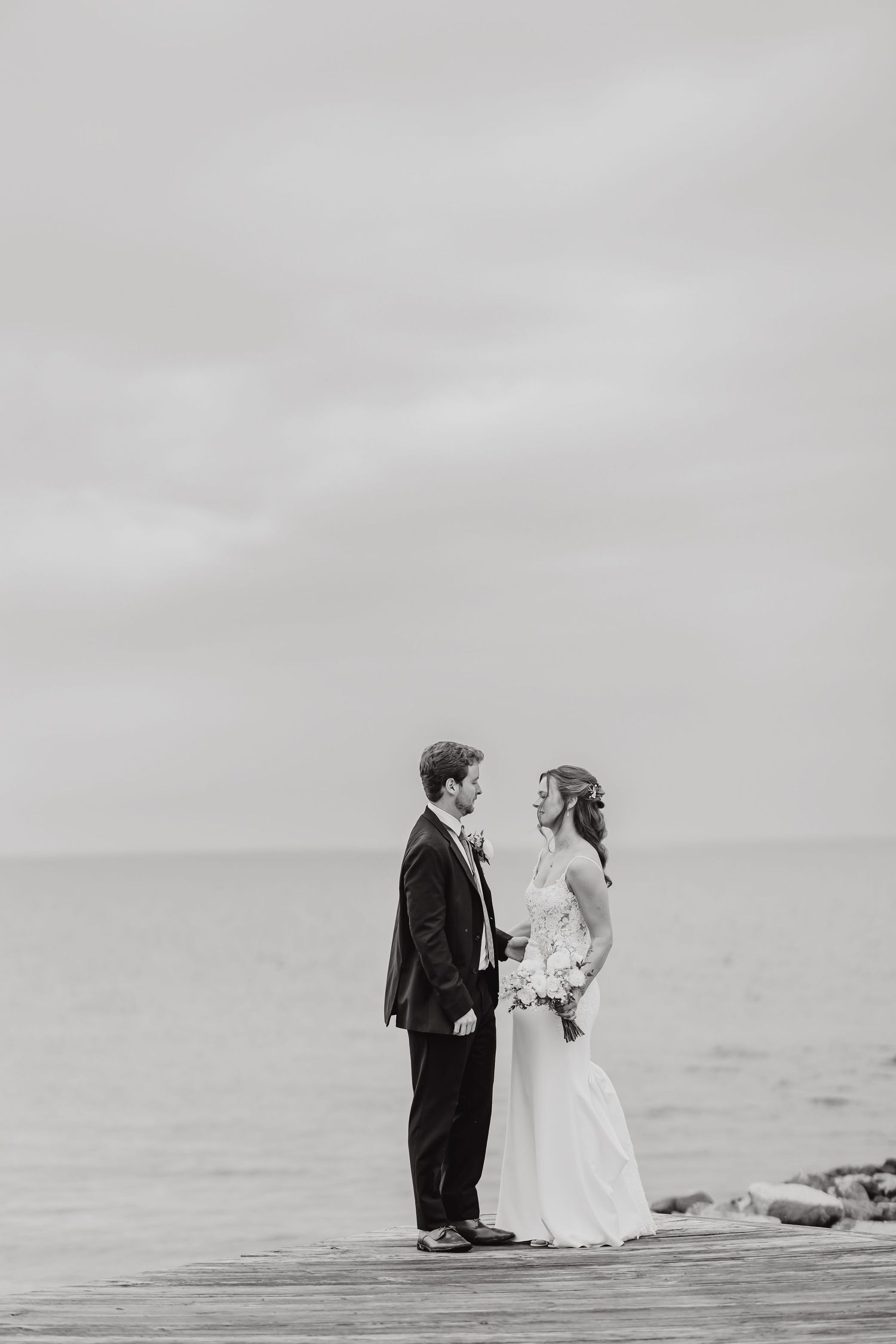 Black and white wedding photo of a couple standing on a wooden pier by the ocean.