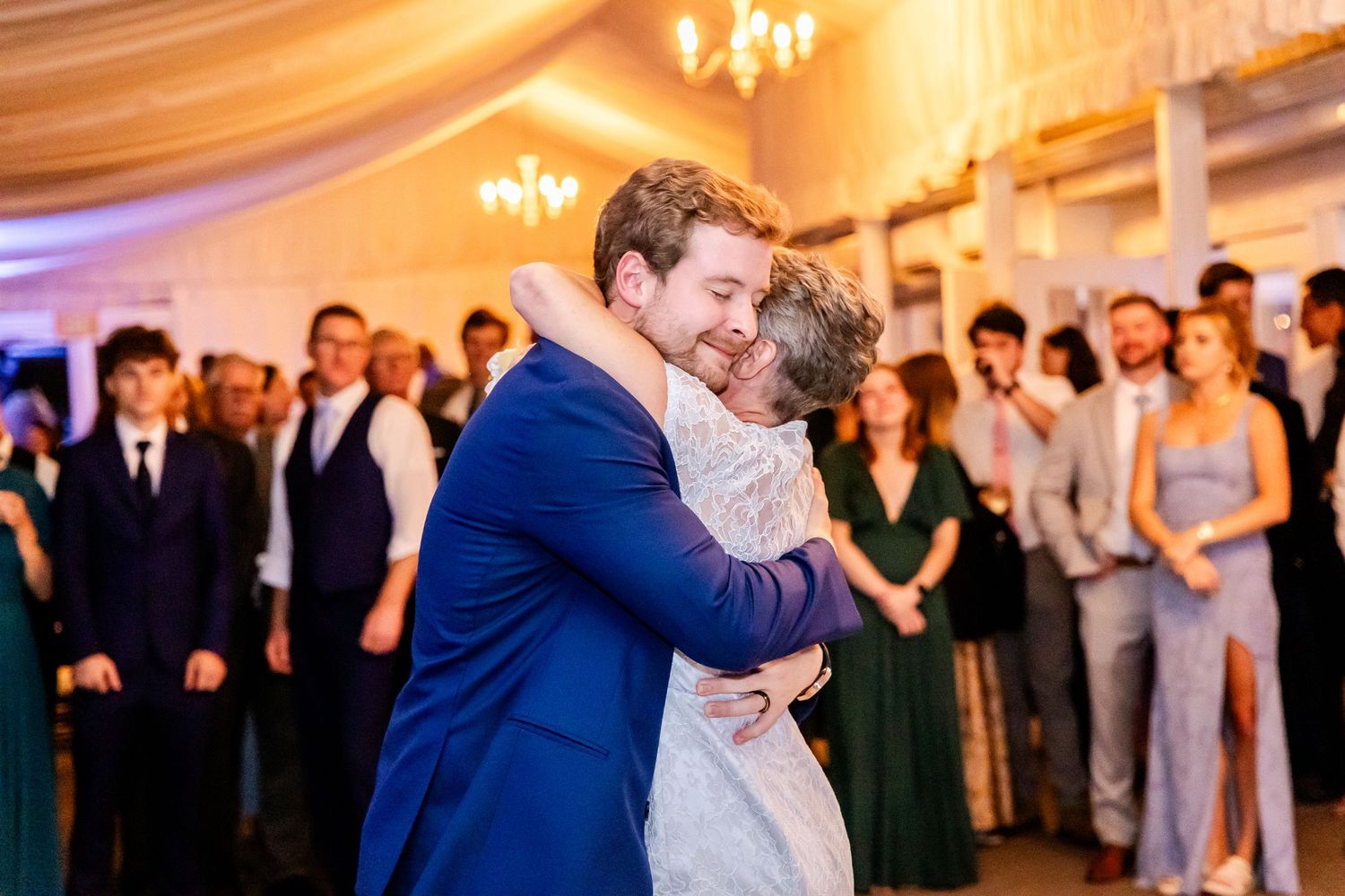 Wedding celebration in a warmly lit ballroom with guests dancing and embracing.