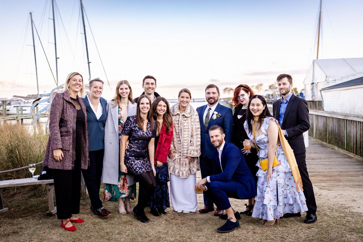 Group of elegantly dressed people posing together on a waterfront boardwalk with sailboats in the background.