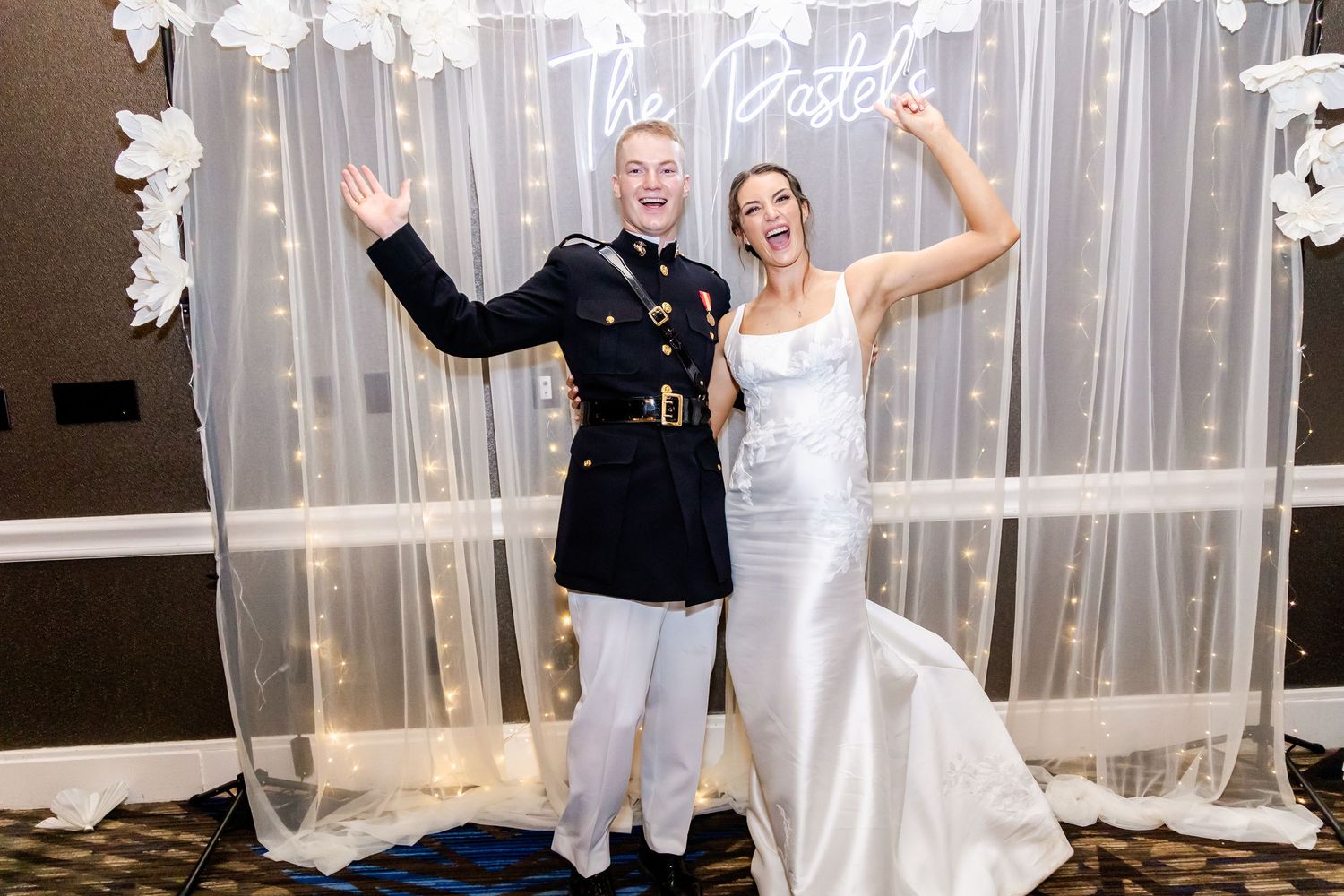 Military groom and bride in white dress pose joyfully against decorative backdrop.
