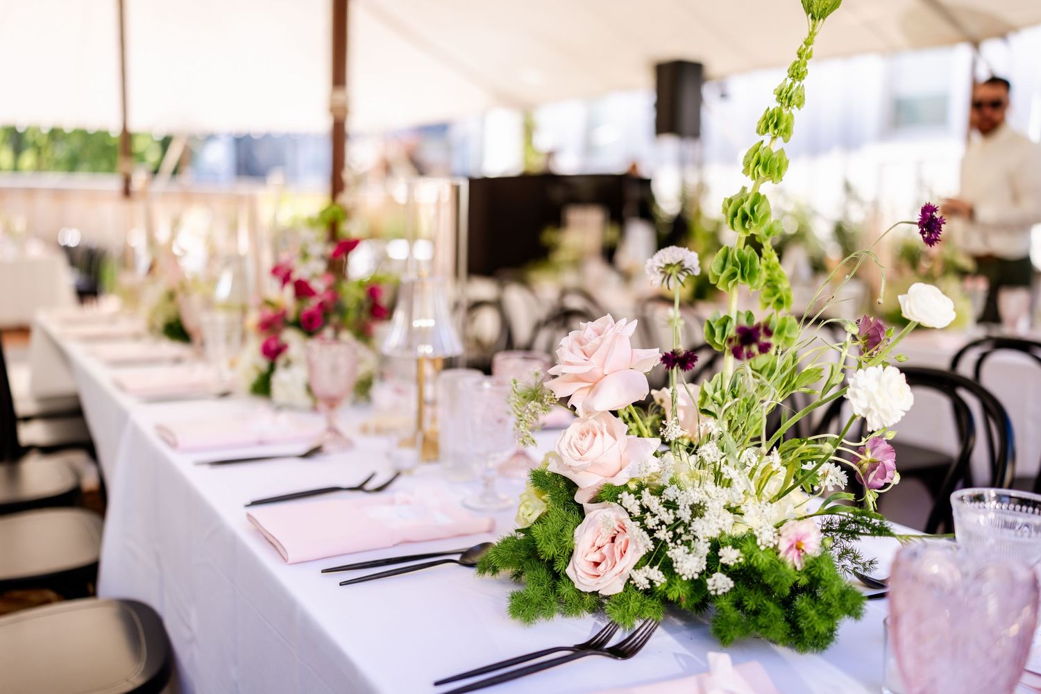 Elegant wedding reception table settings with blush pink flowers and greenery in a bright indoor venue.