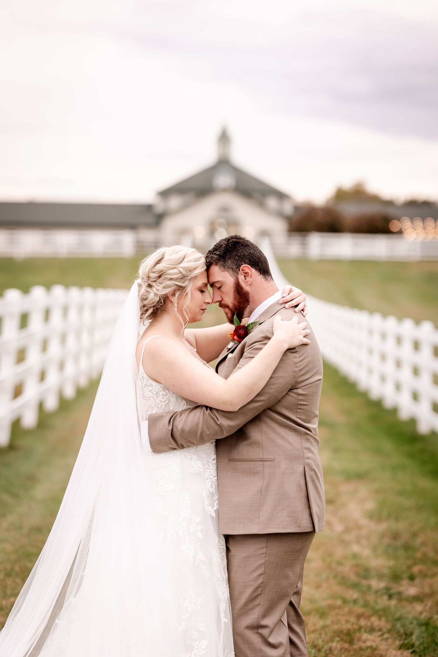 A couple in wedding attire share an intimate moment together in front of a white fence at a rustic outdoor venue.