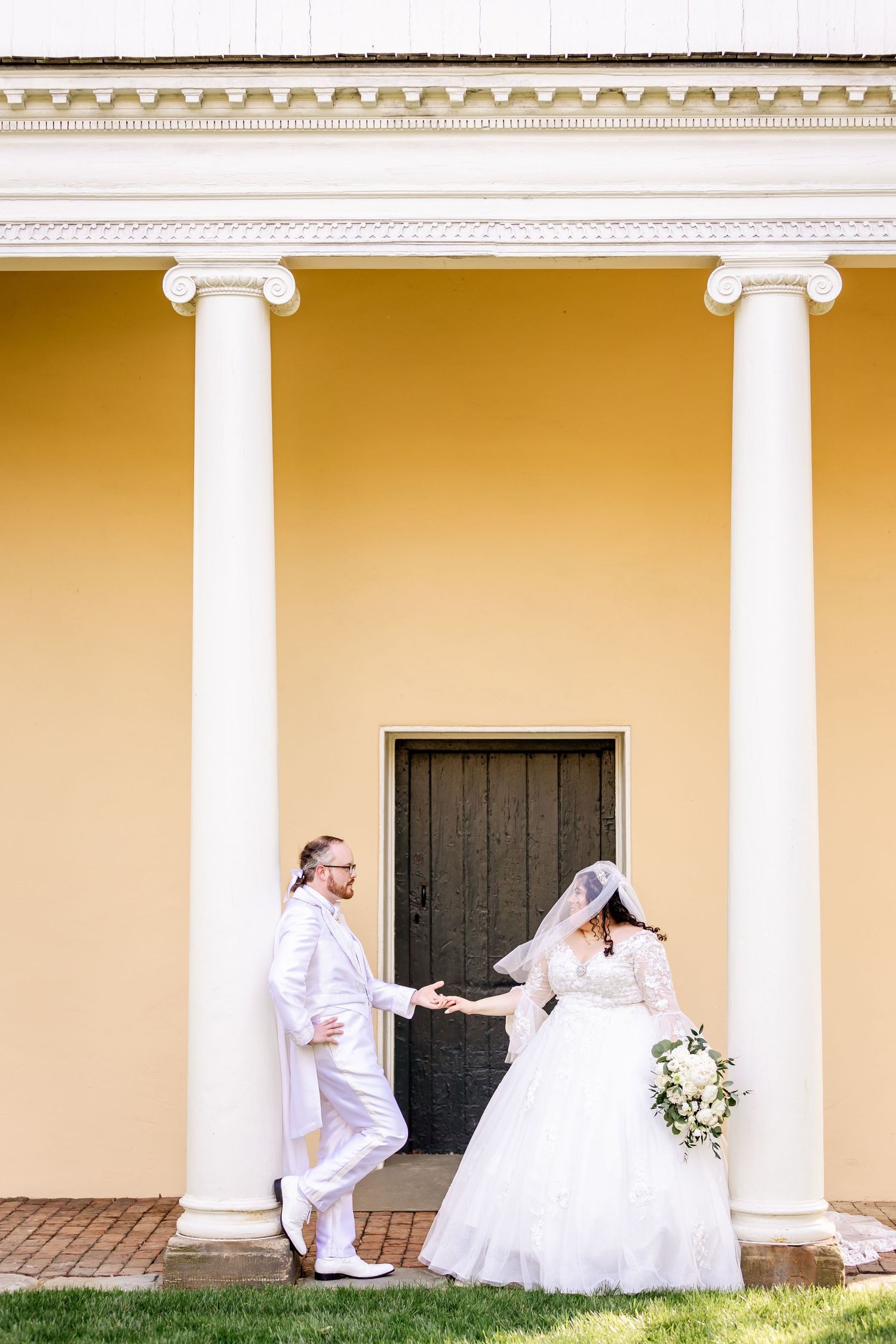 Newlyweds share a romantic moment between white columns of a yellow neoclassical building.