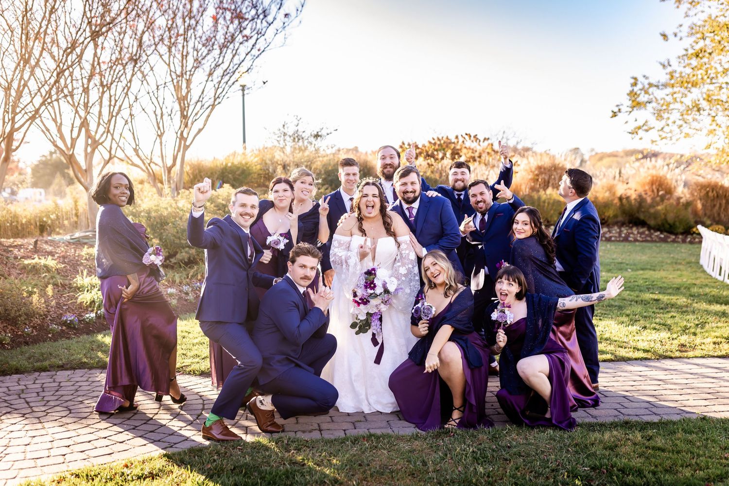 Large wedding party in navy and purple attire poses together outdoors for a fun group photo.