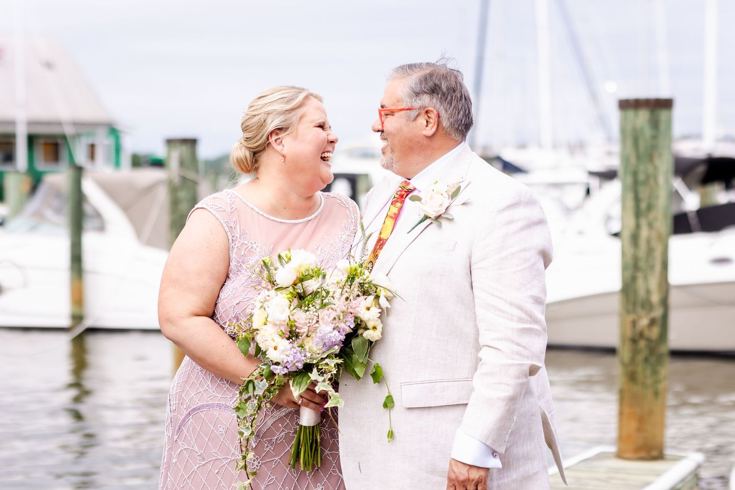 A romantic couple embraces at a marina, dressed in elegant pink and white with a cascading bouquet.