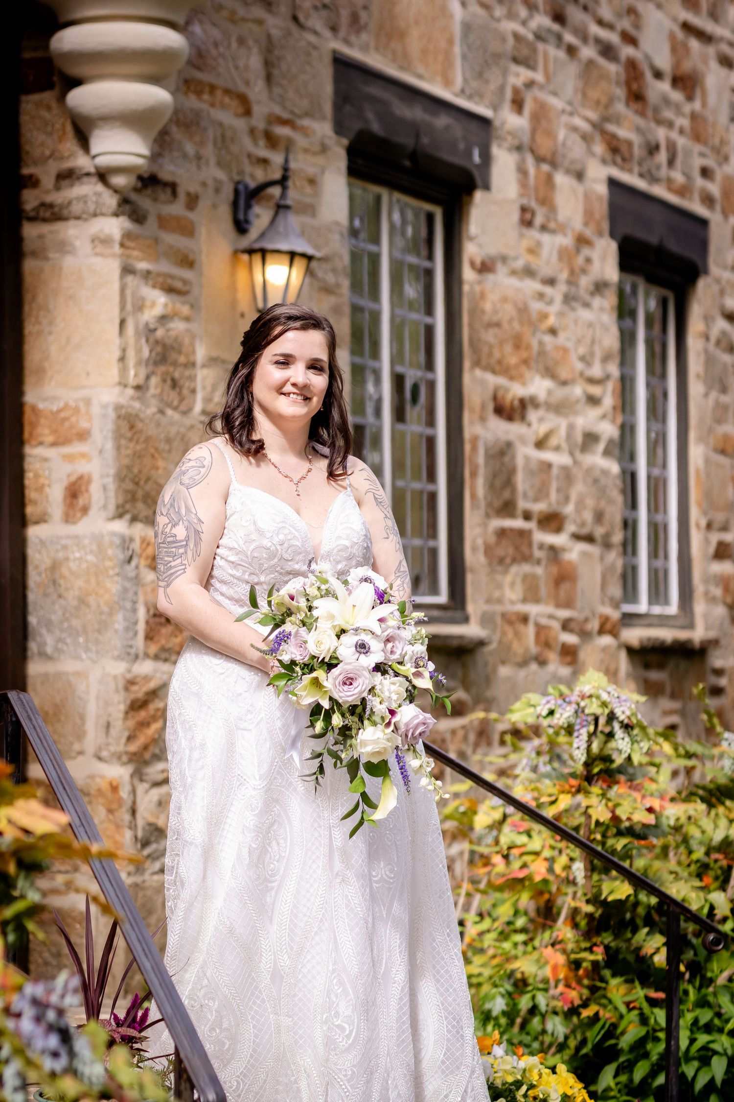 Person in white lace wedding dress holds bouquet outside stone building.