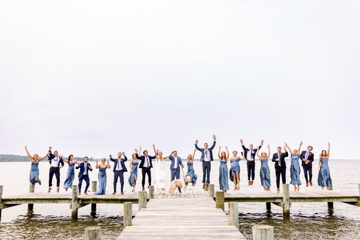 A group of people jumping with raised arms on a wooden pier over a lake during a cloudy day.