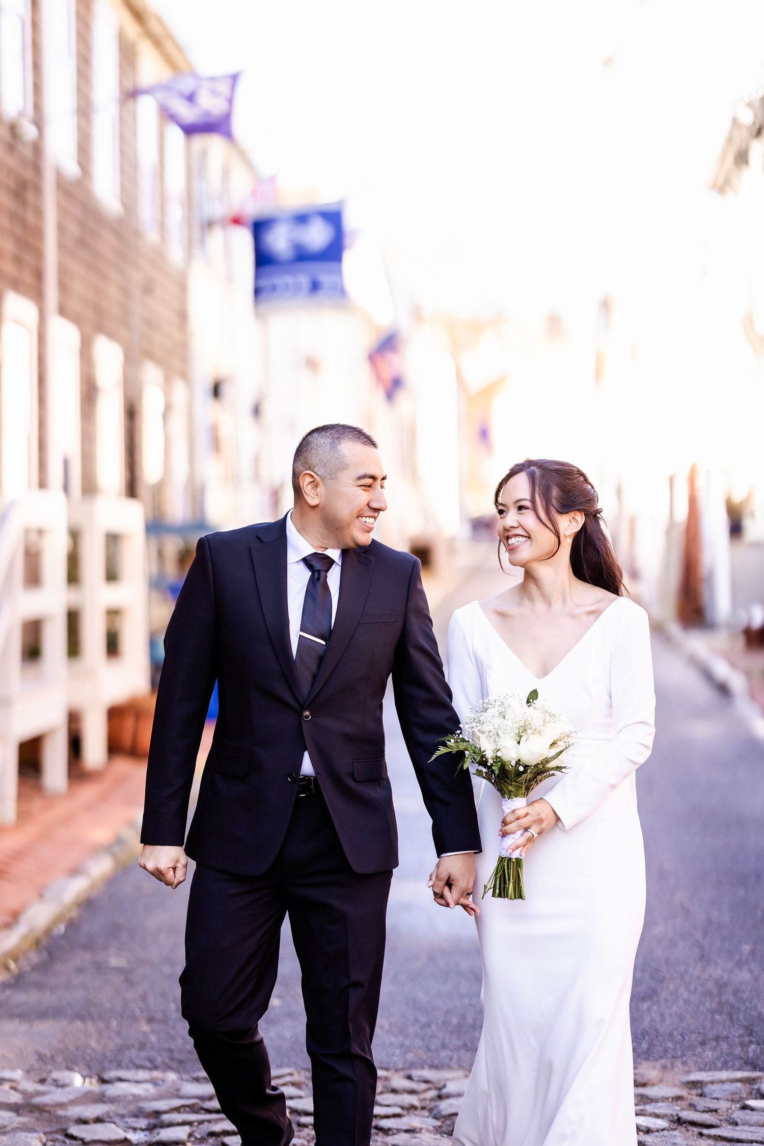 A couple walks down a cobblestone street on their wedding day, with the bride holding a white bouquet.
