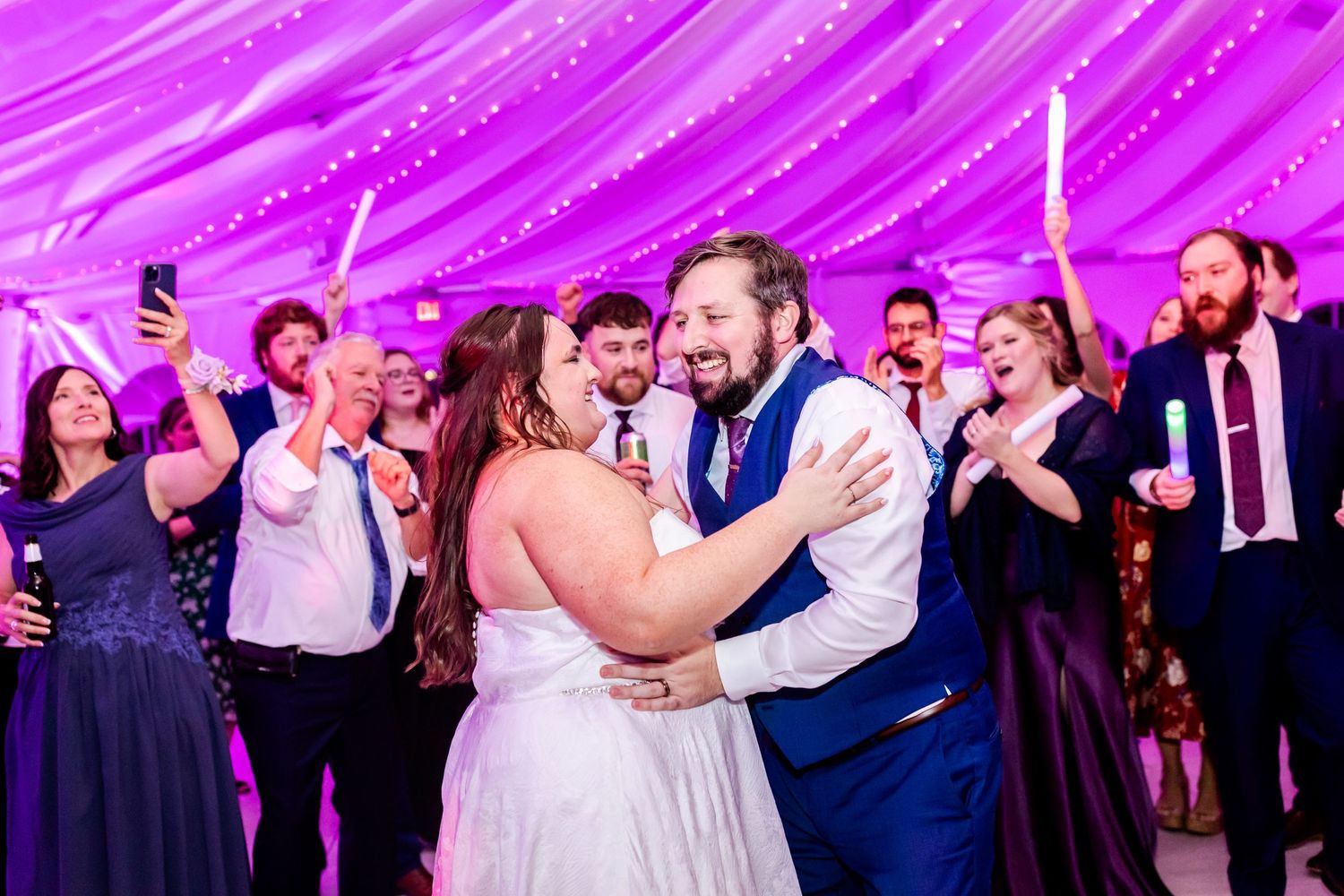A couple shares a romantic dance moment surrounded by guests under magenta draped ceiling.